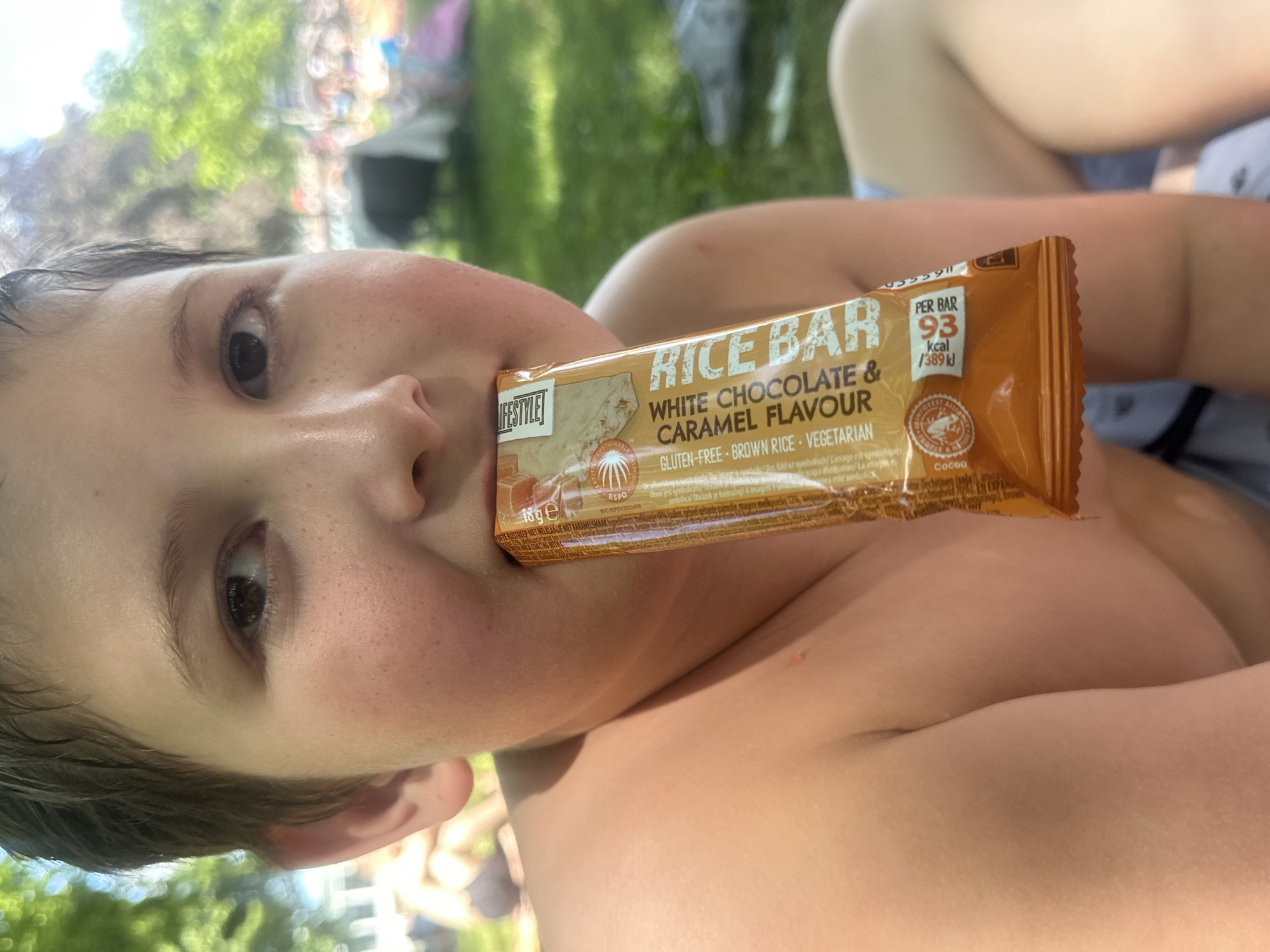 This is a close-up, rotated, outdoor shot of a young person holding a "Lifestyle Rice Bar" with "White Chocolate & Caramel Flavour" in their mouth. The bar is orange and gold and prominently displays nutritional information, stating "PER BAR 93 kcal / 389 kJ". The background is blurred but appears to be a grassy area with dappled sunlight, suggesting it is daytime. The person's face is partially visible, with their eyes open and looking towards the camera. They appear to be relaxed and enjoying the snack. The bar also states it is "GLUTEN-FREE - BROWN RICE - VEGETARIAN". The overall impression is a casual, sunny day outdoor scene with a focus on the snack.