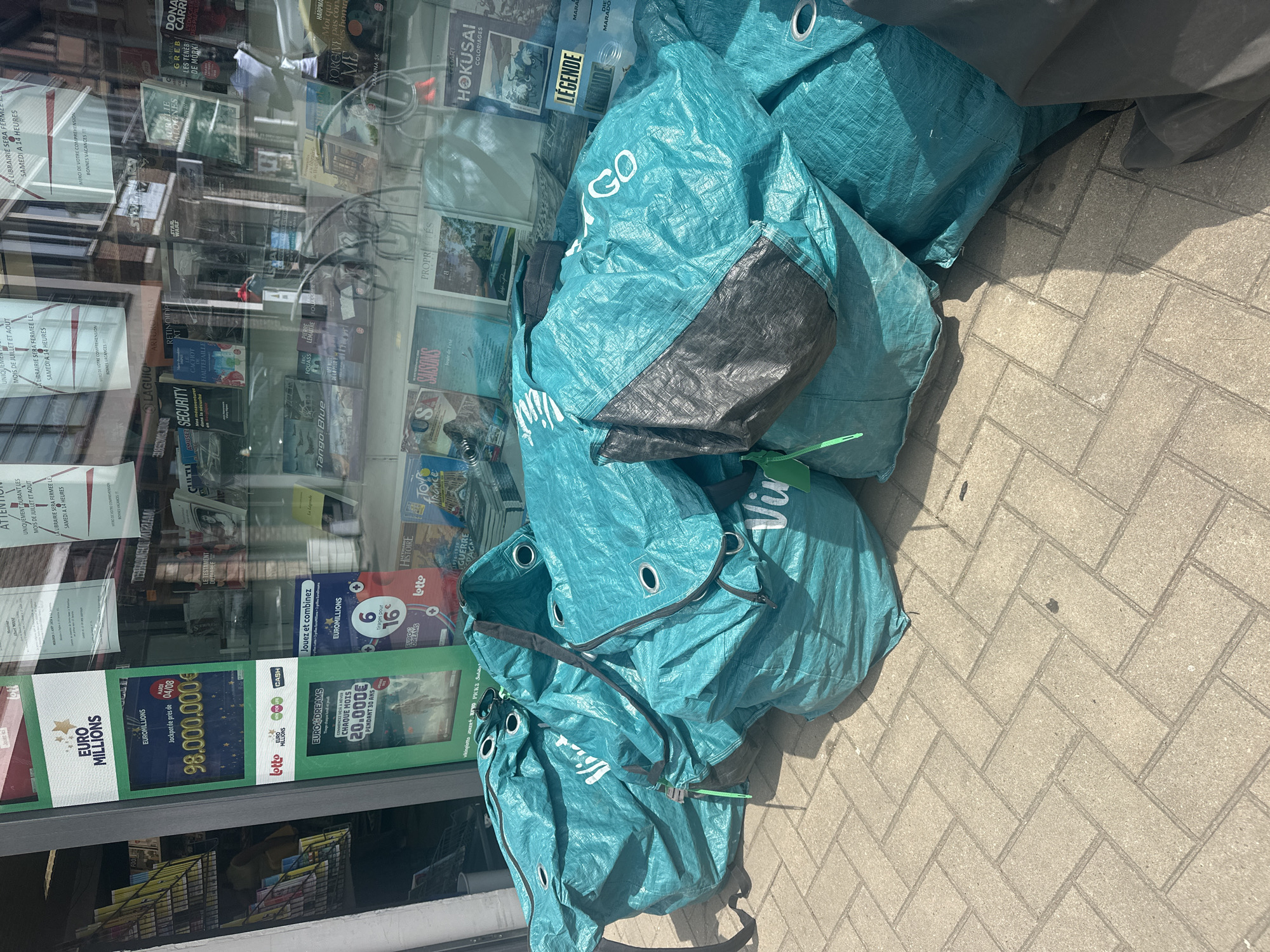 The image depicts a street-level view of a shop window displaying lottery advertisements and various books. Outside the shop, a pile of large, teal-colored tarp bags with black accents and white lettering is stacked on a paved sidewalk. The bags appear to be for waste disposal or storage, with visible drawstrings and grommets. In the background, through the shop window, shelves are stocked with magazines, books, and lottery-related promotional materials, including signs for "EURO MILLIONS" and "EURO DREAMS." Reflections in the window show buildings and sky, suggesting an outdoor urban environment. The lighting suggests it is daytime, with shadows cast by the bags and unseen objects to the right.
