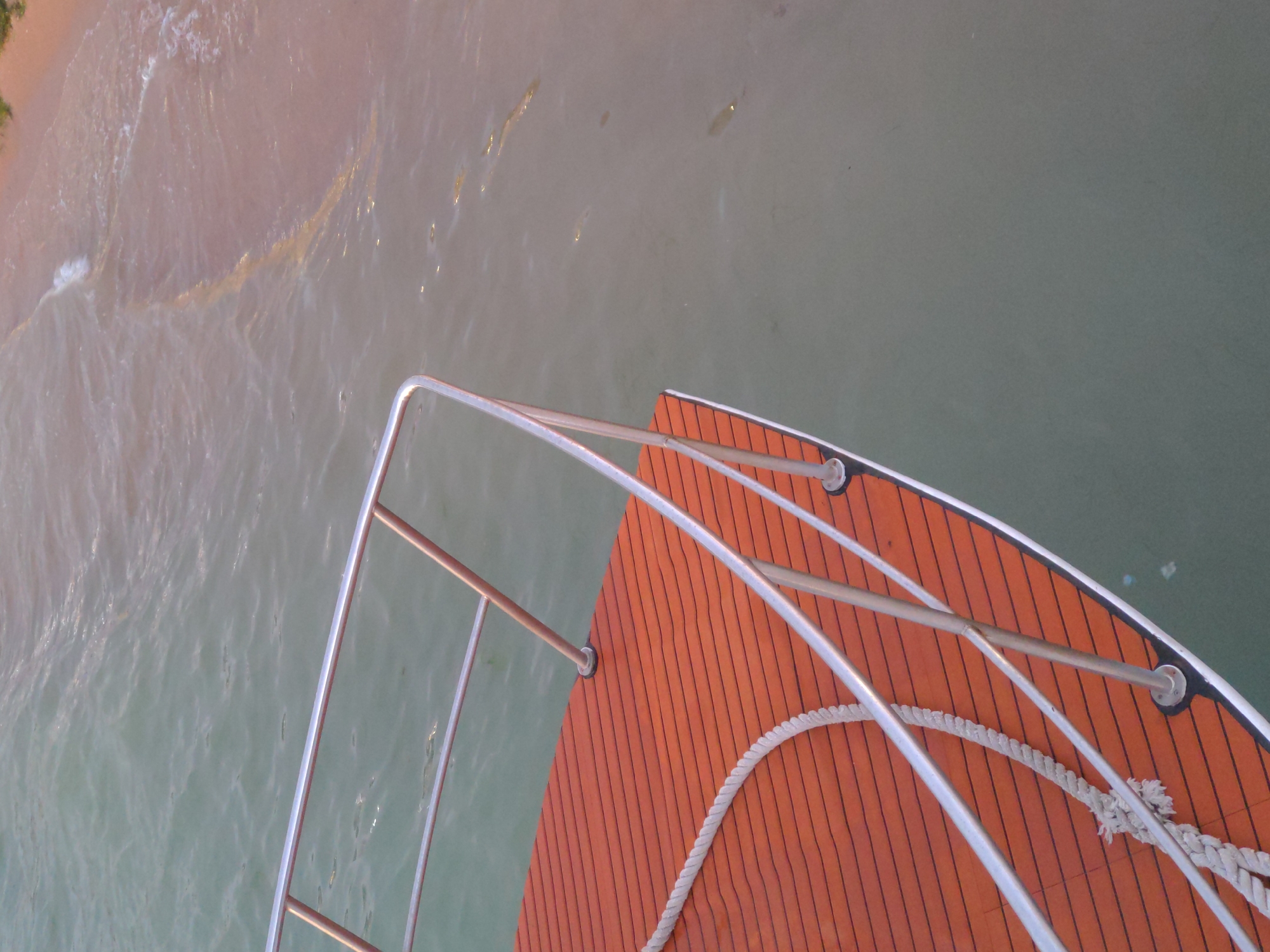 An elevated, close-up shot captures the stern of a boat docked near a sandy shoreline. The boat's deck, made of reddish-brown wooden planks with black lines, is partially visible. A thick white rope is coiled on the deck, and a stainless steel railing with vertical supports runs along the edge. The calm, greenish water surrounds the boat. In the background, a shallow shoreline with pinkish sand is visible, where gentle waves meet the beach. The sky appears overcast, casting a soft light on the scene. The location is the city of Gelendzhik, Russia, as indicated by the context.