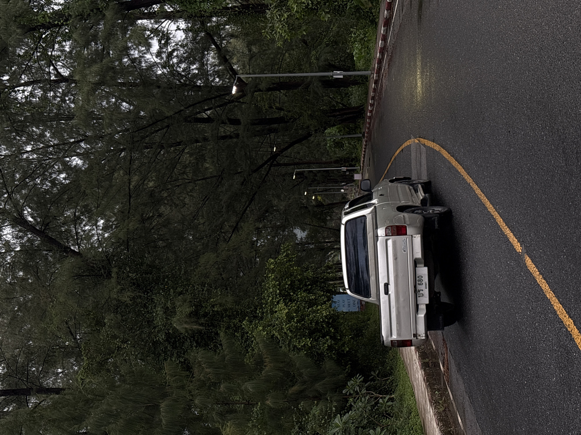 A silver pickup truck is parked on the right side of a wet asphalt road, its tires slightly over the yellow line. The truck's license plate displays Thai characters and the numbers "680". A red and white striped curb runs along the edge of the road. On the left side of the road, a concrete sidewalk is visible, lined with lush green foliage and trees. In the background, a dense forest of tall trees with dark green foliage creates a somber atmosphere. The sky is overcast, contributing to the subdued lighting of the scene. A blue sign is posted on the left, with white text reading "STAY PLAY MEET DINE". The road curves to the right, disappearing into the distance. Streetlights are intermittently placed along the right side of the road.