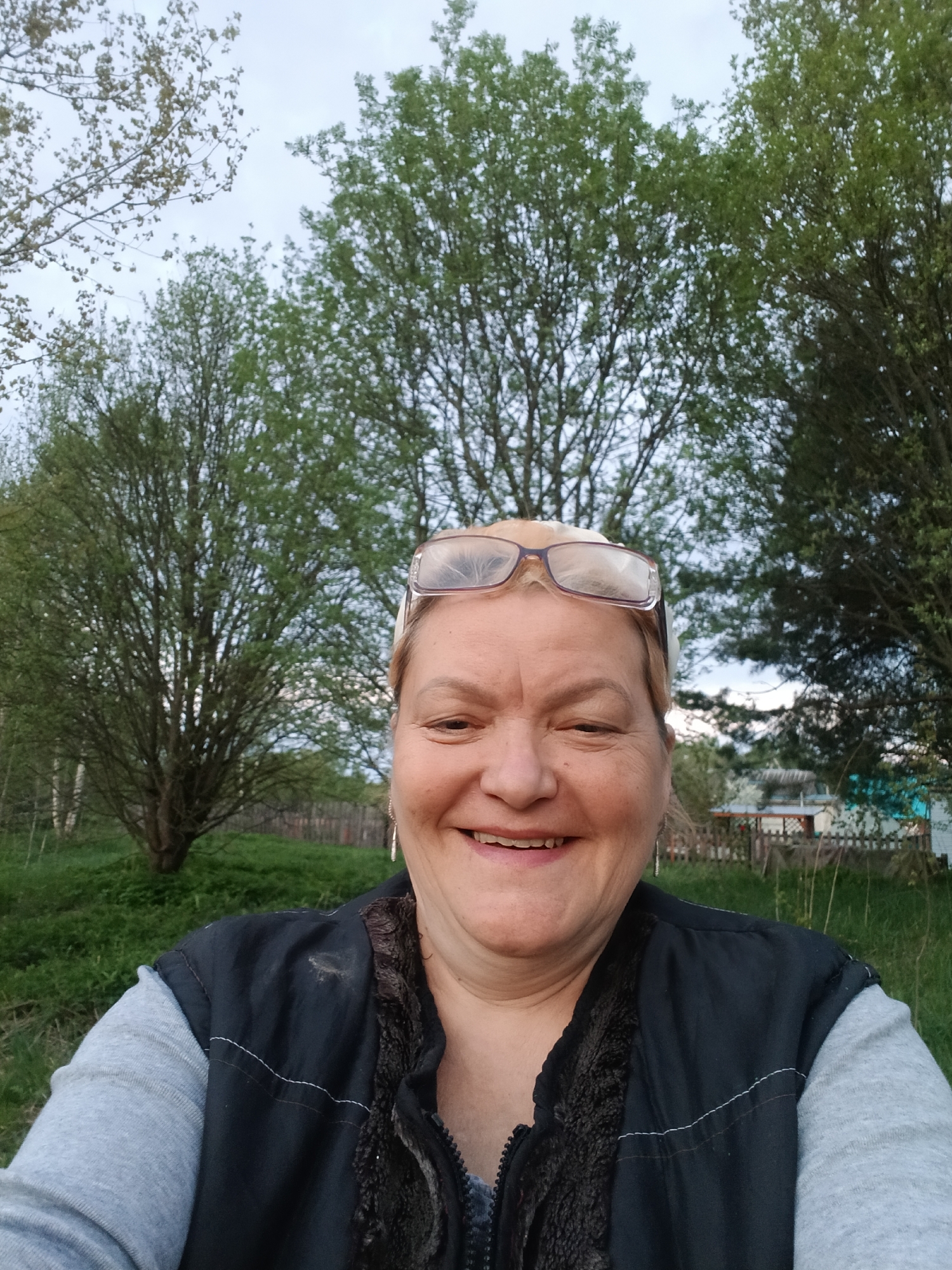 A woman is the main subject of a selfie taken outdoors. She has fair skin, light blonde hair, and is smiling with glasses perched on her head. She is wearing a gray, long-sleeved shirt under a black vest that appears to have a fur-like lining. A wooden fence and a small building can be seen in the background behind her on the right, surrounded by trees and grass. This scene is in Красный Маяк, Russia.