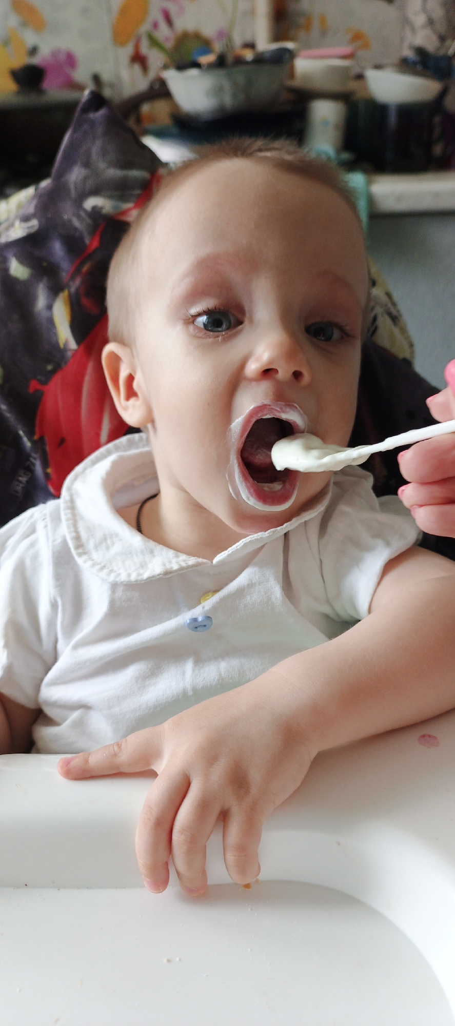 A close-up, vertical photograph shows a baby being fed indoors. The baby, with fair skin, light hair, and wide blue eyes, is seated in a white high chair, looking intently towards the person feeding them. Their mouth is open wide, revealing a pink tongue, and is smeared with a white, creamy food. A white spoon, also laden with the white substance, is being held to their mouth by an adult hand with pink nail polish, visible on the right side of the frame.
The baby wears a white collared shirt with short sleeves, featuring one yellow and one blue button. A thin, dark necklace or cord is visible around their neck. Their right hand rests on the white high chair tray, which has a few crumbs and a faint pink smudge.
The background, out of focus, shows a colorful, floral-patterned fabric or wallpaper behind the baby's head and shoulder. Further back, various household items such as bowls and mugs are visible on a surface, suggesting a domestic setting like a kitchen or dining area. The lighting indicates it is daytime. There is no clearly discernible text visible in the image.