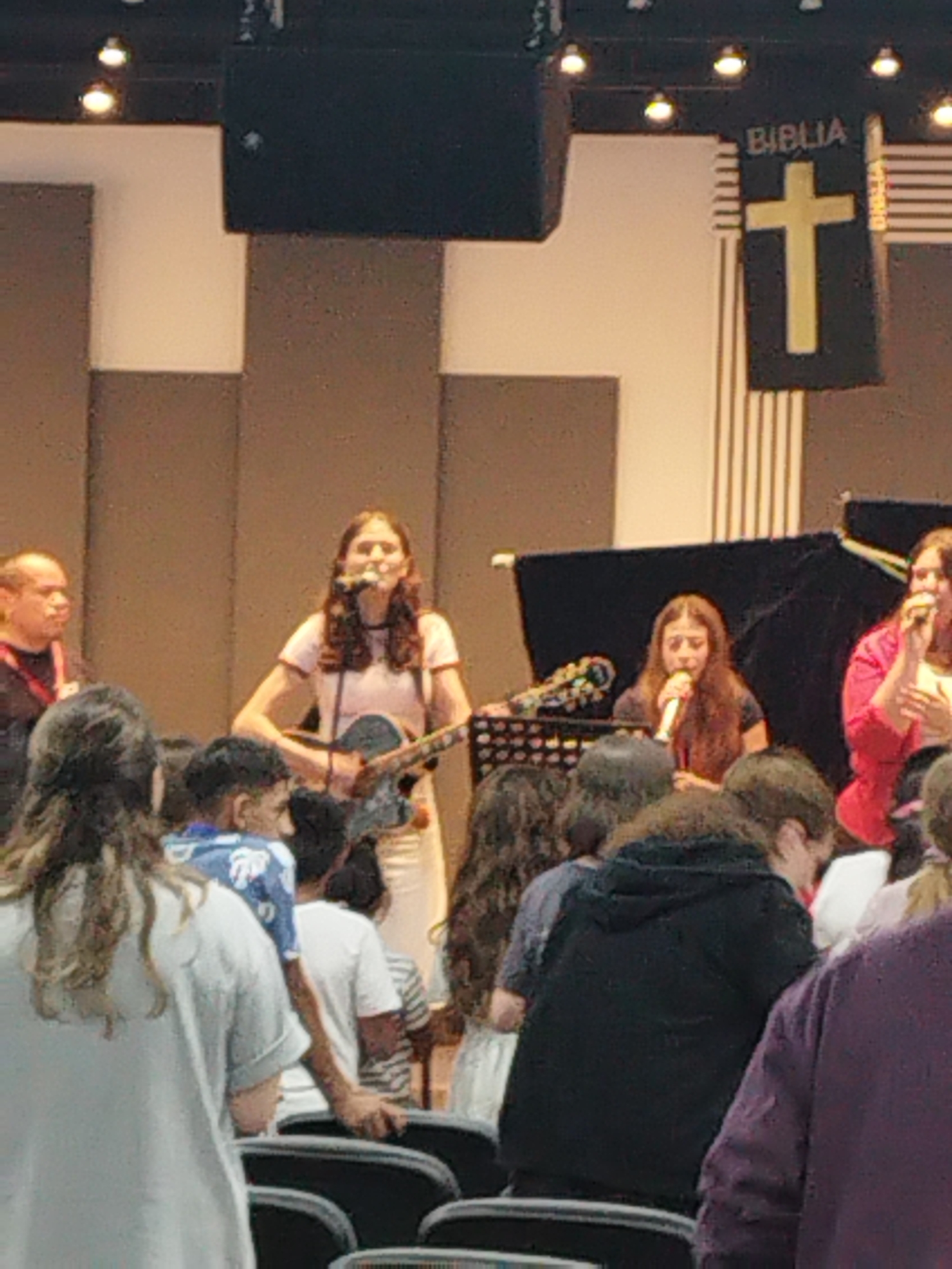 The image depicts a worship service, likely taking place in a church or a similar venue in Bucharest, Romania. In the foreground, several individuals are seated, with their backs to the camera, suggesting they are part of the congregation. The middle ground features a group of people performing on a stage. A young woman in the center is singing and playing an acoustic guitar, wearing a pink t-shirt and white pants. To her right, another young woman is singing into a microphone, wearing a pink blazer. Further to the right, a woman in a red top is also singing. Behind the performers, a black curtain is visible. To the left of the stage, a man in a black shirt and lanyard is standing. In the background, a banner with a cross and the word "BIBLIA" (Bible) is displayed. The environment appears to be an indoor setting with stage lighting and acoustic paneling on the walls. The overall atmosphere suggests a religious gathering or concert.