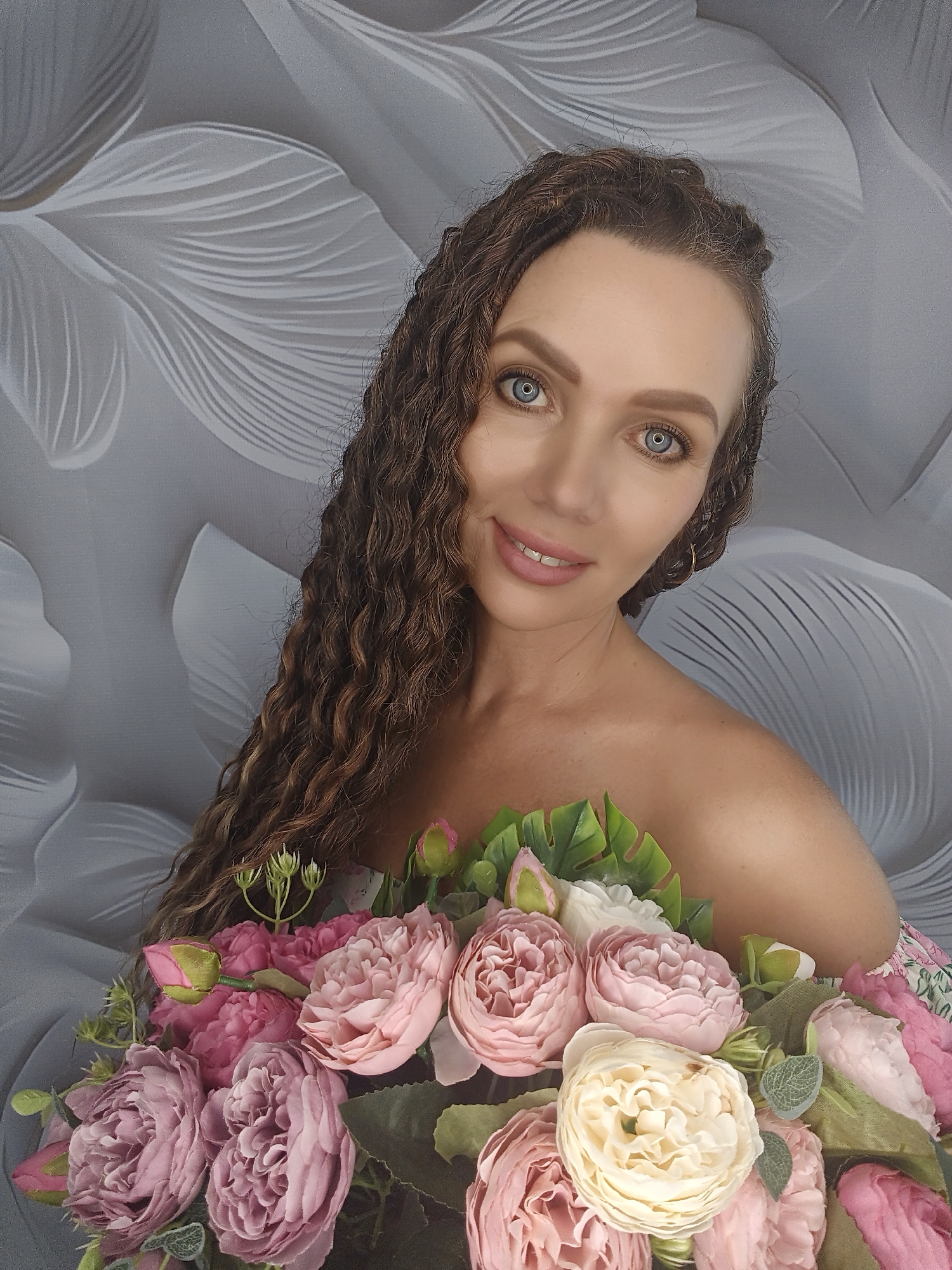 A medium shot captures a woman with long, curly brown hair and blue eyes from an eye-level perspective. She is positioned in front of a grey, textured background that resembles abstract floral patterns. The woman is bare-shouldered and holding a large bouquet of artificial flowers, primarily in shades of pink, cream, and green. The bouquet obscures her torso and appears to be part of her attire. Her head is tilted slightly to her right, and she is looking directly at the camera with a subtle smile. The lighting is soft and even, highlighting her facial features and the details of the flowers.