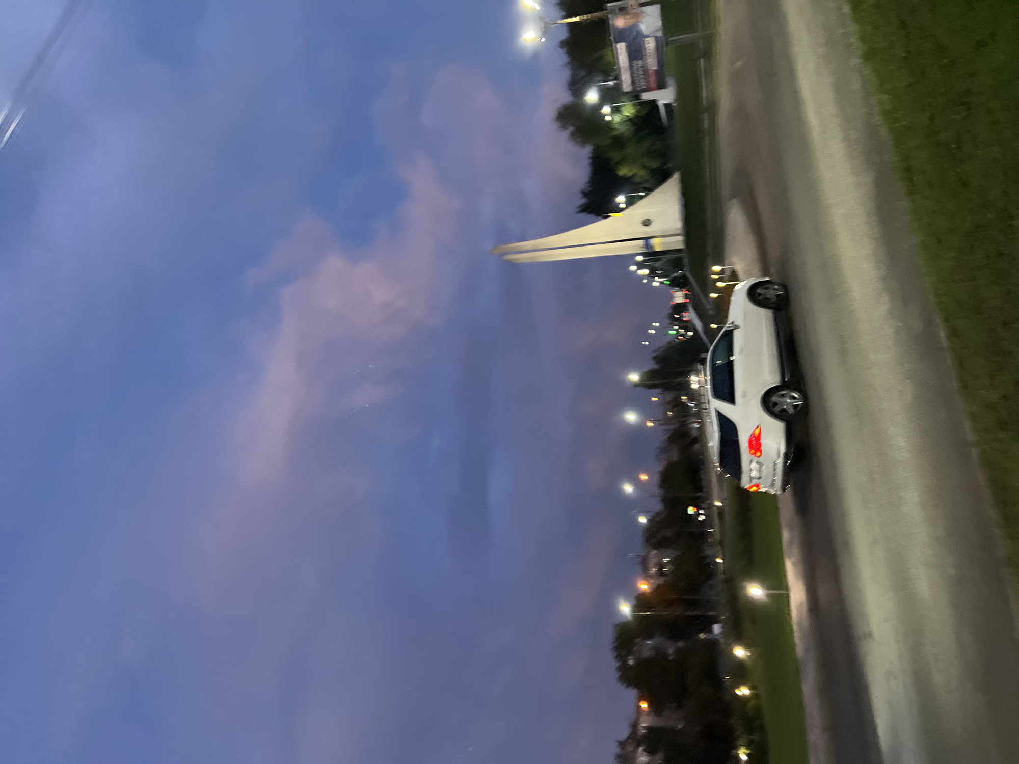 In the city of Unknown, Unknown, a white Mercedes-Benz SUV is captured in motion, driving on a paved road. The vehicle's brake lights are illuminated, suggesting it is slowing down or stopped. In the background, a large, white, modern sculpture stands prominently against a twilight sky. The sky is a deep blue with soft hues of pink and purple, indicating either sunrise or sunset. Streetlights are visible, casting a warm glow on the scene, interspersed with trees and buildings. A billboard is also present on the right side of the frame, partially obscured by trees. The overall scene conveys a sense of urban environment at dusk, with elements of modern architecture and natural light.
