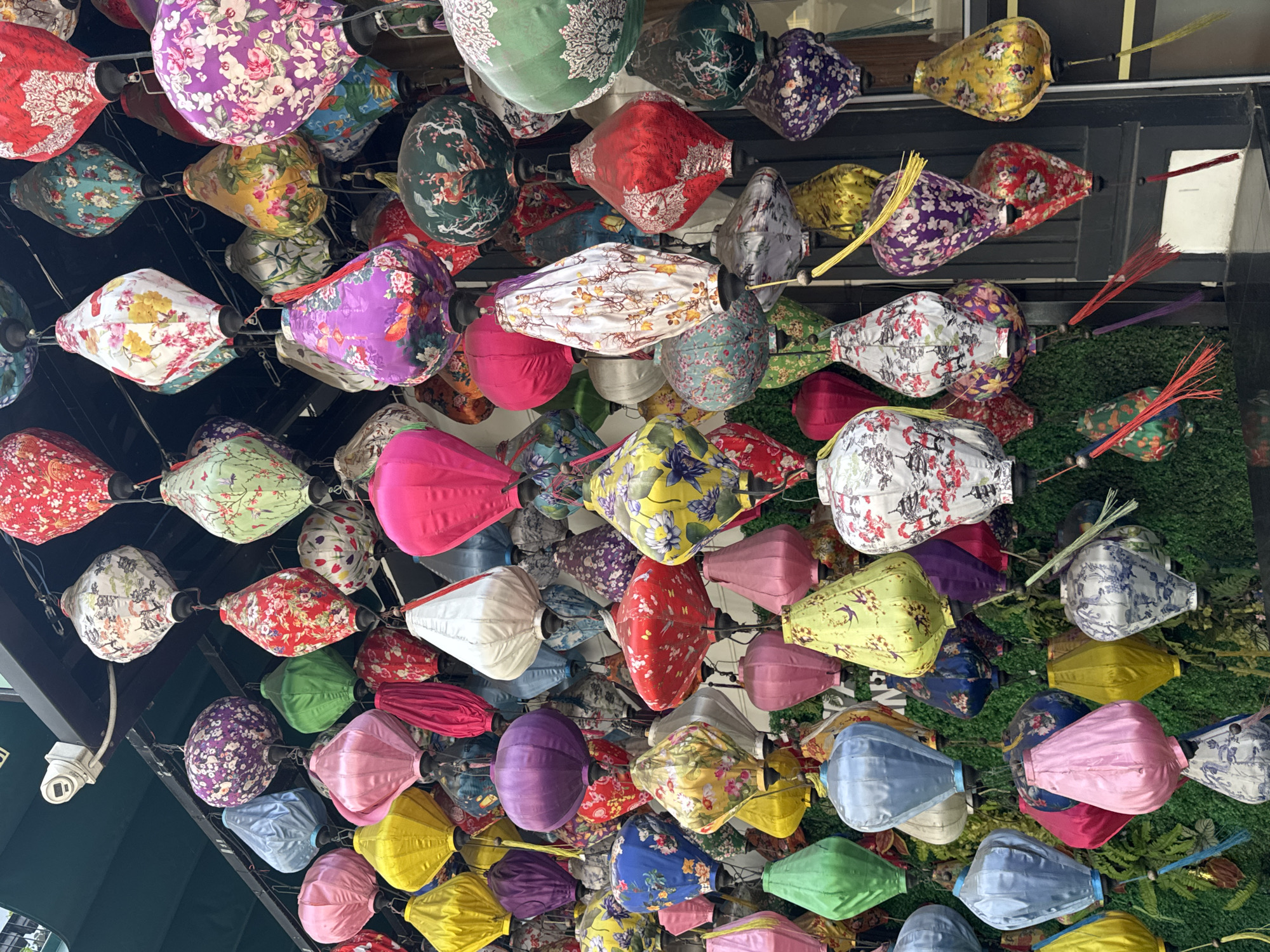 A vibrant array of colorful Vietnamese lanterns hangs from the ceiling, filling the frame. The lanterns are teardrop-shaped with rounded bottoms and come in a variety of colors and floral patterns, including pink, purple, yellow, green, red, and blue. Some have tassels attached. The lanterns hang from black ceiling fixtures and are illuminated from above. In the background, a green wall can be seen, and a white security camera is visible on the left side of the image. The scene is likely located in the city of Đà Nẵng, Vietnam, as this city is known for its colorful lanterns.