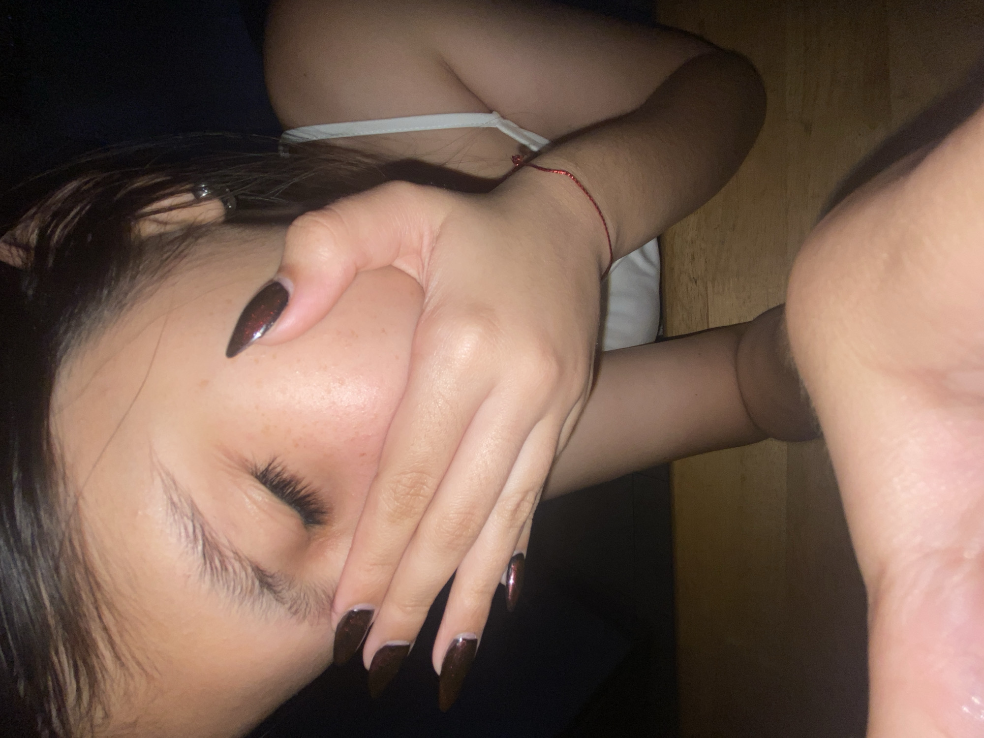 A close-up indoor shot presents a person with dark hair and brown stiletto nails, their face partially obscured by their hand. The person wears a white tank top, with a red string bracelet visible on their wrist. The background includes a dark surface and a light brown wooden surface. The person's eye is closed. There is no information to suggest that this image was taken in Cotoperíz, Venezuela.