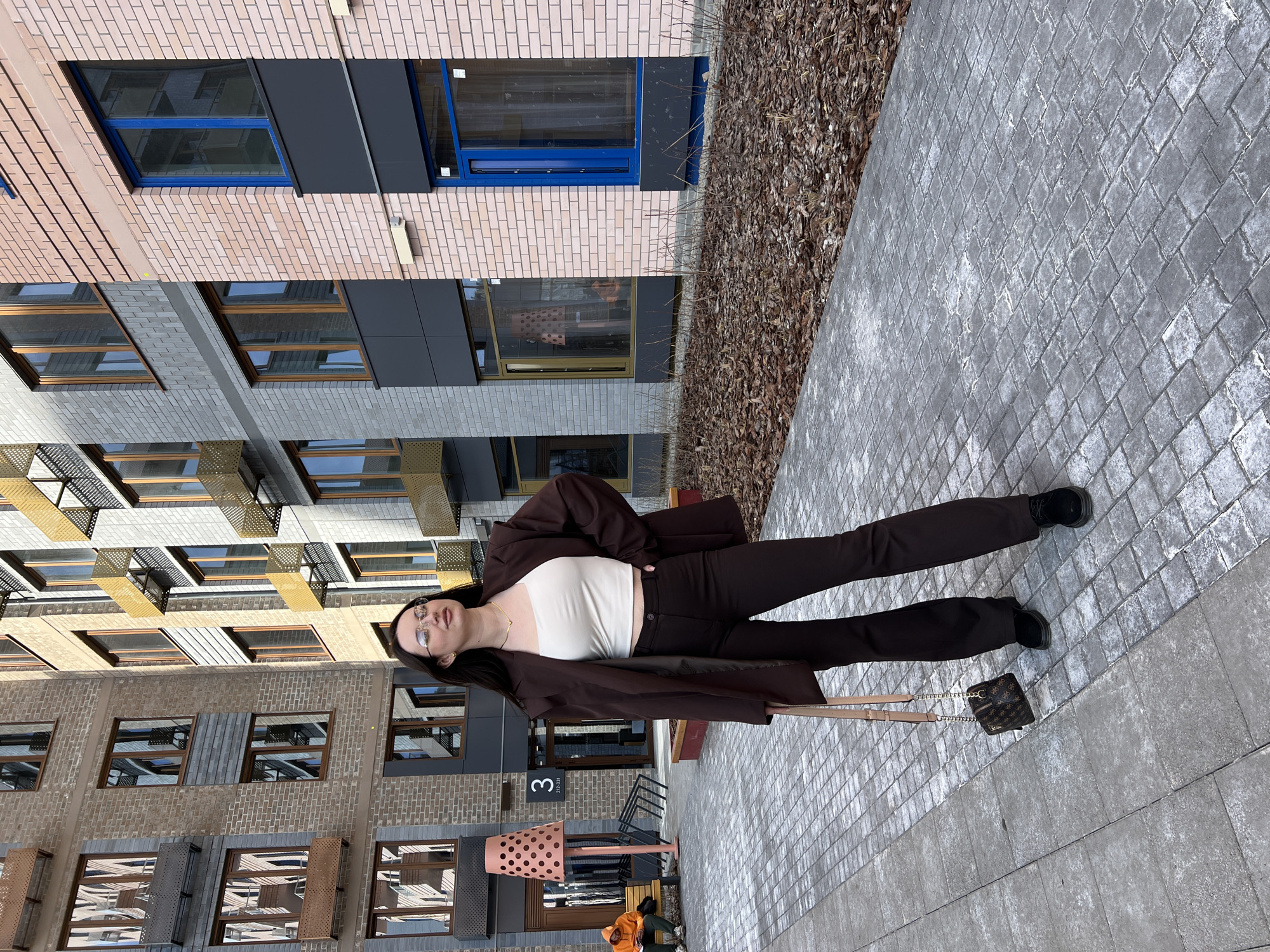 The image shows a woman lying on a gray brick sidewalk in an urban environment. She is wearing a brown pantsuit over a white top and black shoes. A small brown handbag rests next to her. Behind her is a modern building with a mix of brick and dark gray siding. The building features windows with brown and blue frames. The number "3" is visible on the building's exterior. Across from the sidewalk is a patch of brown mulch or leaves. The city is Tyumen, Russia.