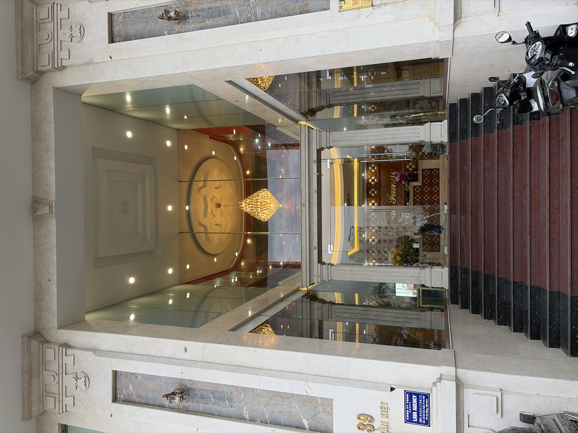 The image shows the entrance of a building in Da Nang, Vietnam, identified by a sign for "LAVA AGENCY" with an address that includes "TP. Da Nang, Viet Nam". The building has a grand facade with marble columns and decorative elements. A set of red and black granite stairs leads up to the glass entrance doors. Inside, a large crystal chandelier is visible, along with a decorated ceiling and interior lighting. Two motorcycles are parked at the bottom of the stairs. A sign on the left side of the building displays the number "39" and Vietnamese text.