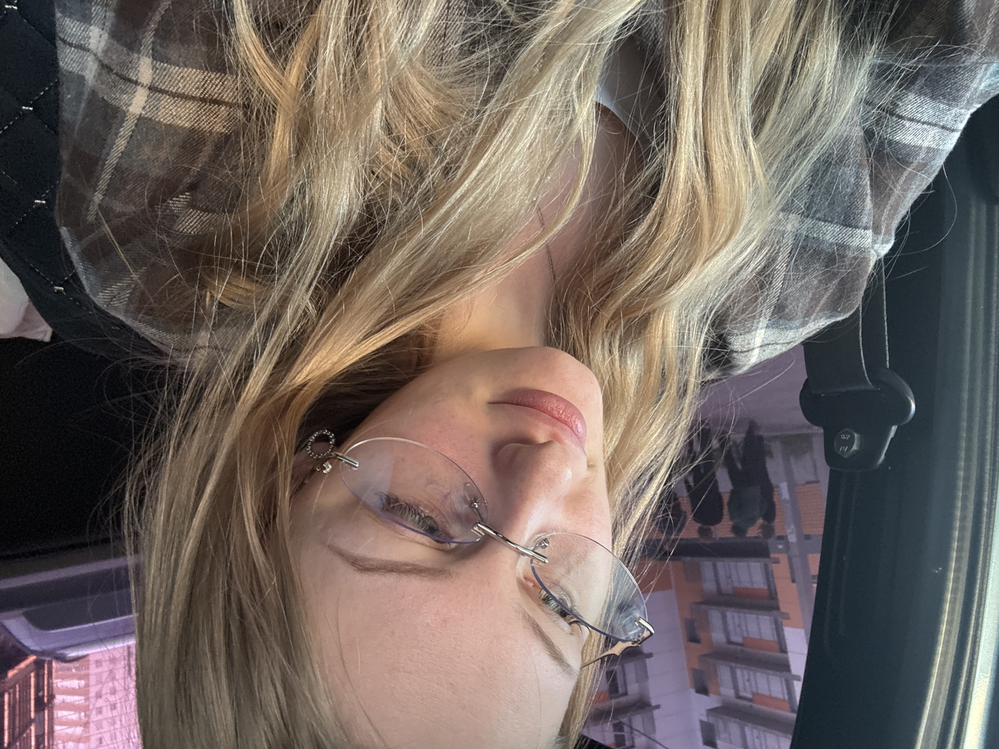 A young woman is seen in a close-up shot, positioned upside down relative to the frame. She has fair skin, blonde hair, and is wearing frameless eyeglasses. She appears to be inside a vehicle, as indicated by a seatbelt buckle and the glimpse of a building visible through the window. The building's exterior has an orange and white color scheme, characteristic of the architecture in Perm, Russia. She is wearing a plaid flannel shirt. Her hair is long and flowing.