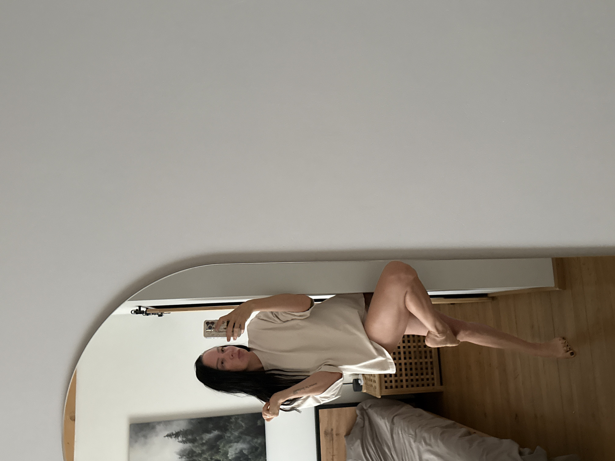 A full-body mirror selfie is taken by a woman with long, dark hair in a bedroom. The woman is wearing a loose-fitting, cream-colored oversized t-shirt that extends past her thighs. Her right leg is bent and crossed over her left, with her right foot resting on her left calf. Her left leg is extended downwards, with her left foot bare and slightly elevated off the wooden floor. She is holding a smartphone in her right hand, angled to capture the reflection. Her left hand is raised to her head, with her fingers casually resting on her hair.
In the background, reflected in the mirror, is a bed with gray linens. To the left of the bed is a wooden nightstand with a woven front. Behind the nightstand, a large framed artwork depicting a misty forest scene hangs on the wall. The room features light-colored walls and a wooden floor. The setting appears to be a residential interior, possibly in Chaikovsky, Russia, given the context provided.