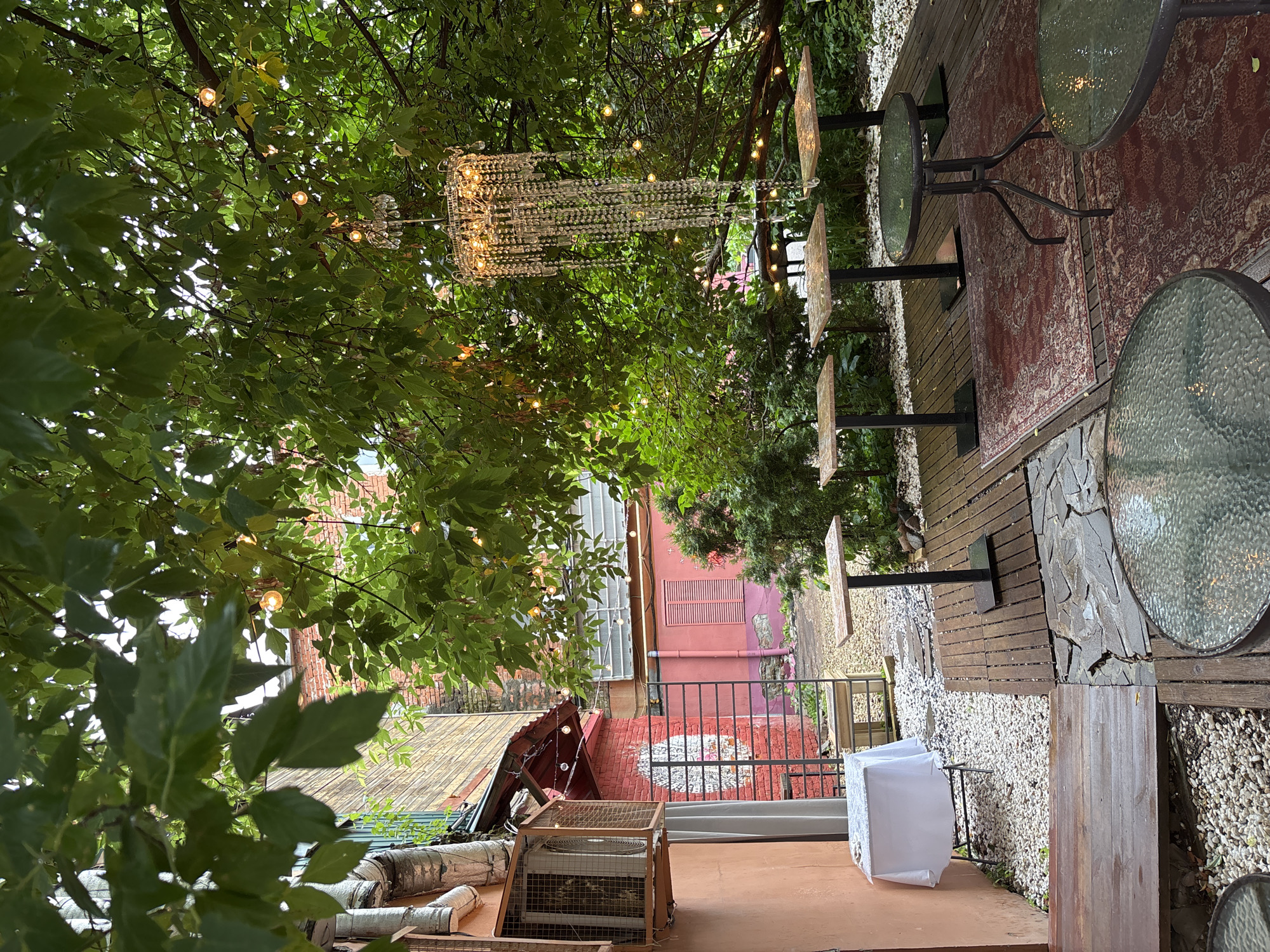 The image depicts an outdoor seating area of a cafe or restaurant in Nizhny Novgorod, Russia. The area is partially covered by the lush green foliage of a tree, from which a crystal chandelier and string lights are suspended. There are several tables of varying sizes and designs: a small white-clothed table near a building, four tall square-top tables arranged in a row on a wooden deck, and two round glass-top tables, one prominently in the foreground. A patterned rug is placed on the wooden deck between the tall tables and the round tables. The background reveals brick buildings with windows and pipes, and a pale pink wall with shutters. The ground is a mix of wooden decking, gravel, and stone paving. The overall impression is of a charming, possibly bohemian, outdoor dining space.