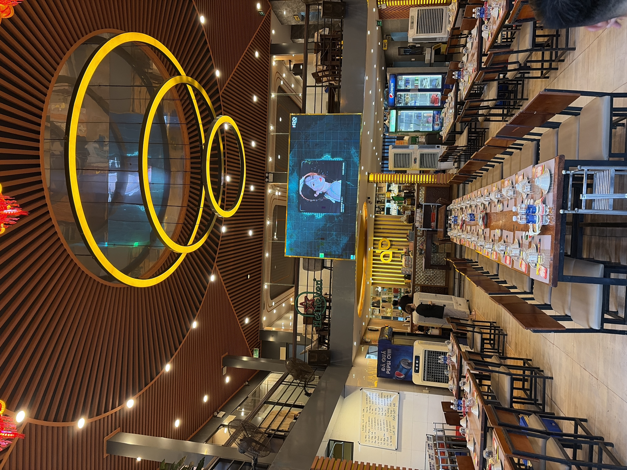 This image depicts the interior of a restaurant in Đà Nẵng, Vietnam. The restaurant features a modern design with a high ceiling adorned with intricate wooden slats and circular yellow pendant lights. Large screens display graphics, including an animated portrait of a woman. Dining tables are set with place settings for multiple diners, and numerous chairs are arranged around them. In the background, a bar area is visible with shelves stocked with bottles. Several large fans and air conditioning units are present in the space, and a Pepsi refrigerator is prominently displayed. The overall atmosphere suggests a busy dining establishment.