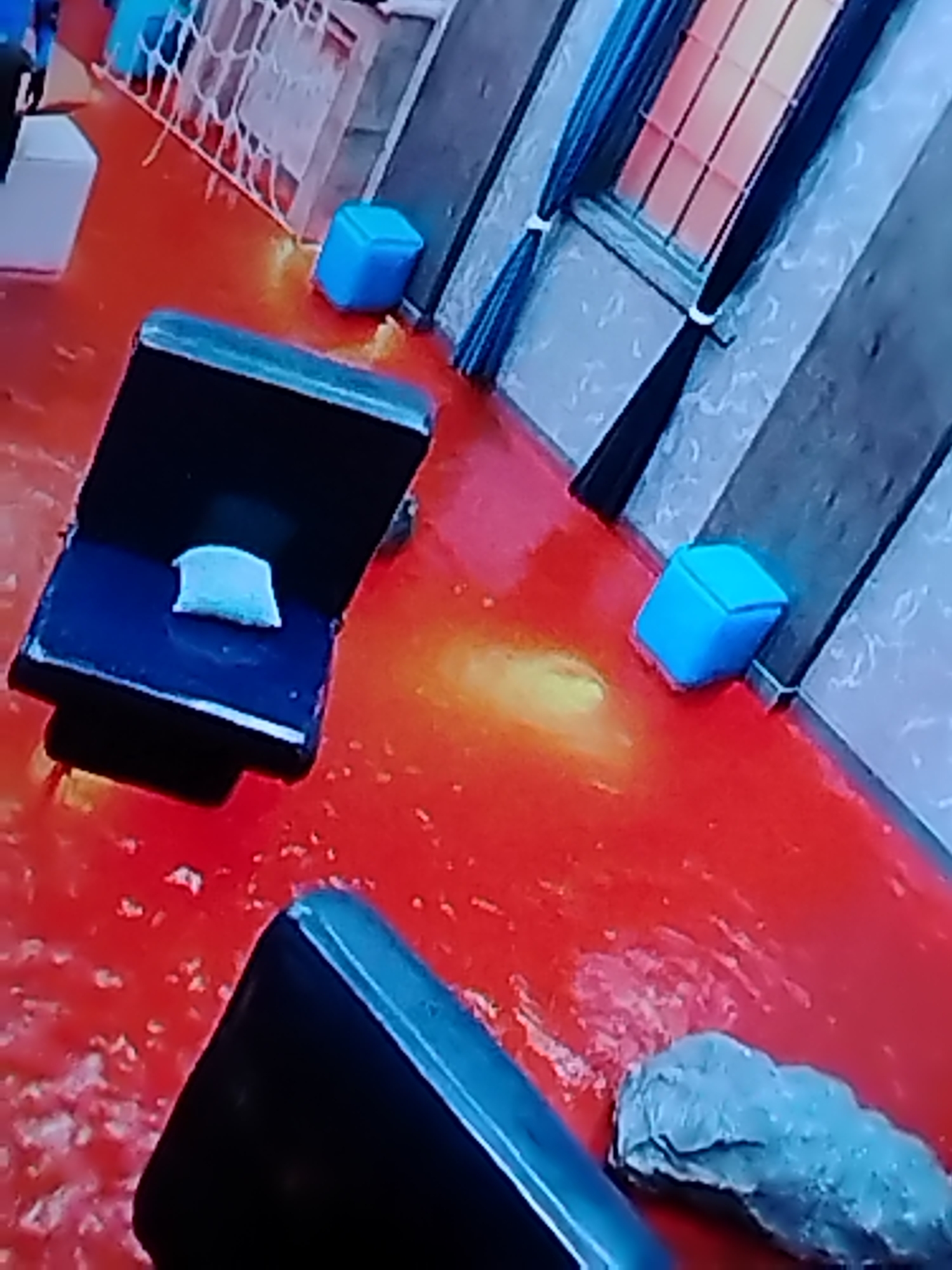 The image shows a room with a red floor, possibly painted or flooded with a red substance. There is a black chair with a white cushion on it, as well as several blue cubic stools scattered around the room. A window with blue curtains is visible in the background. Based on the context provided (Rusanovo, Russia), it can be inferred that this is likely an art installation or a set design. A grey, rock-like structure sits near the bottom right. Another black chair sits in the bottom left.