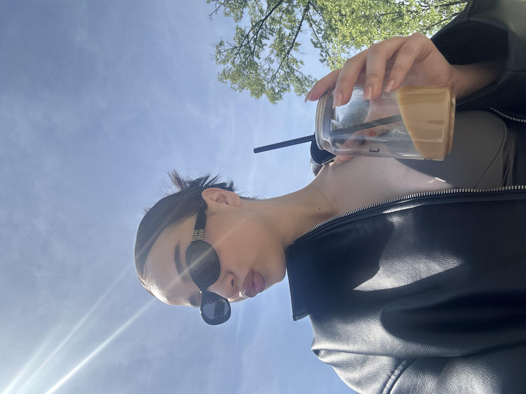 A young woman with dark hair pulled back into a bun is posing with a clear plastic cup containing a tan-colored liquid, likely a coffee drink. She is wearing dark round sunglasses, a light-colored top, a delicate gold necklace, and a black leather jacket. The background is a blue sky with the sun shining and green trees. The photo was likely taken outdoors, in the daytime, in an urban environment. There is no specific information to indicate this image was taken in Palilula Urban Municipality, Serbia.