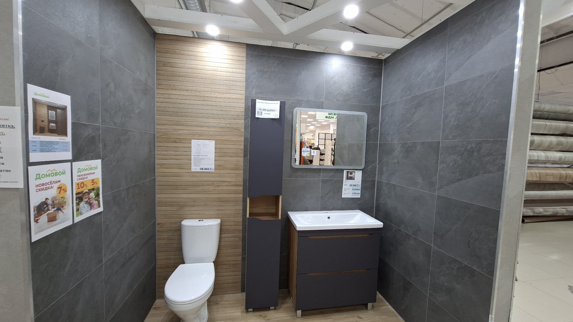 The image captures a staged bathroom display in what appears to be a retail setting, potentially in the city of Chaikovsky, Russia, given the presence of Cyrillic text on signage and product information.
The display features a toilet, a gray storage cabinet with a partially open compartment, a vanity with a sink and drawer storage, and a square mirror. The walls are tiled with large, gray rectangular tiles on two sides and horizontal wood-look planks on the third.
Several informational signs are visible: one next to the toilet lists a price of "10 385", another on the wall indicates discounts for new residents ("НОВОСЁЛАМ СКИДКА!") and senior citizens ("ПЕНСИОНЕРАМ СКИДКА!"), with percentages of 7%, 10%, and 5% respectively. There is also a price for "10 300 руб/шт" (10,300 rubles per piece).
The setting appears to be a home improvement or furniture store, judging from the lighting, product displays visible in the mirror's reflection, and the partial view of stacked rolls of flooring material to the right. The overall impression is of a well-lit retail space presenting a bathroom design concept for customers.