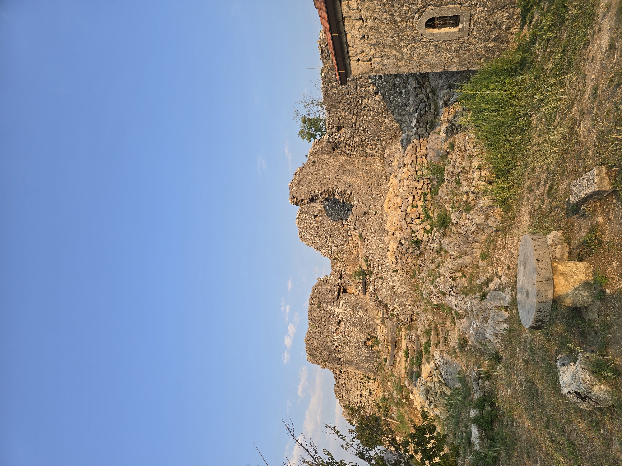 The image captures a view of the Medun fortress ruins in Medun, Montenegro. The scene is bathed in the warm light of a setting sun, casting long shadows across the stone structures and overgrown landscape. The foreground features a rudimentary stone table and bench, weathered by time and elements. To the right, a portion of a stone building with a tiled roof and a small, barred window is visible, likely part of the fortress complex. The main focus is the crumbling stone walls of the ancient fortress, which dominate the middle ground, their rough-hewn stones and irregular shapes speaking of centuries of history. Steps made of stone and wood ascend the slope towards the ruins. The background is a clear, pale blue sky with a few wispy clouds. Green vegetation, including grass and some trees, grows amidst the rocks and ruins, indicating a natural and somewhat untamed environment. The overall impression is one of historical significance and natural beauty, with the ruins standing as a testament to the past against the backdrop of a tranquil sky.