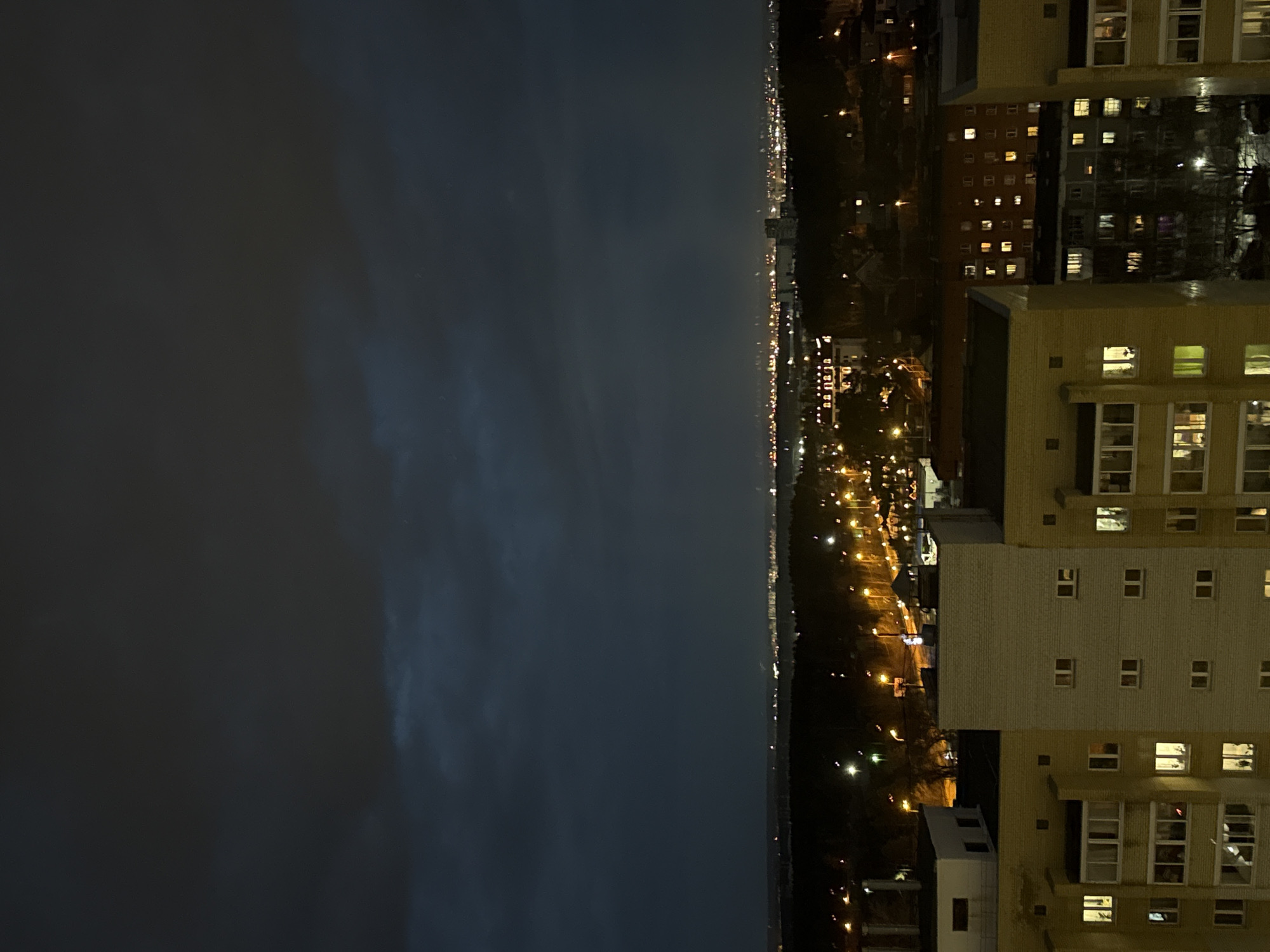 The image shows a night view from a high vantage point, possibly from a building. The right side of the image features a tall, light-colored apartment building with numerous lit windows. The scene overlooks a city area illuminated by streetlights. Beyond that is the dark and somewhat hazy sky. This scene is likely in Yekaterinburg, Russia, given the prompt's context.
