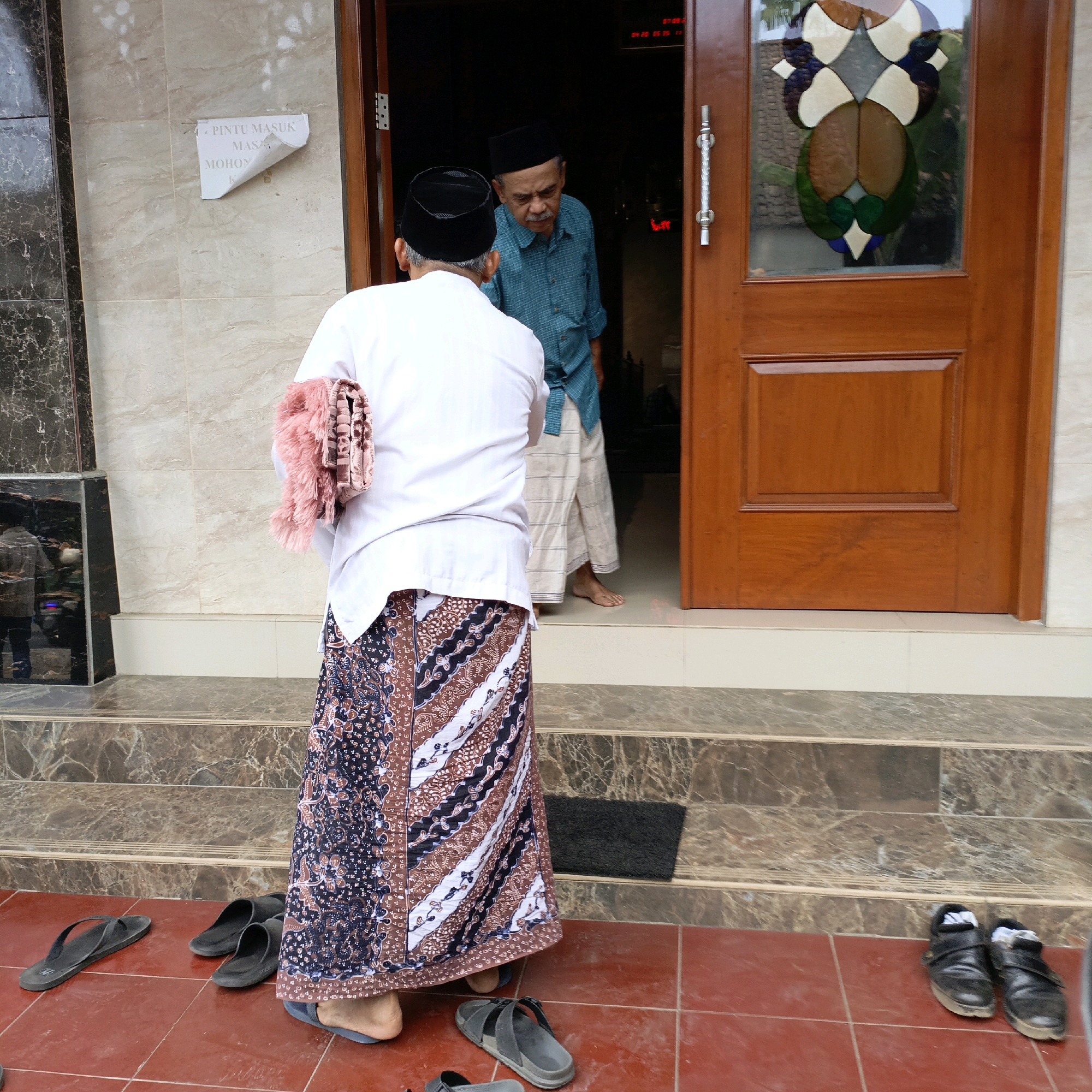 Two men in traditional Islamic attire are standing on the steps of a building in Salatiga, Indonesia, near a wooden door. The man in the foreground, facing away from the camera, wears a white shirt and a patterned sarong. He holds a pink prayer mat over his shoulder. The man in the background, facing the foreground man, wears a teal shirt and a white sarong. He appears to be looking at the other man. A sign on the wall to the left of the door reads "PINTU MASUK MOHON" in Indonesian, indicating a men's entrance. Several pairs of sandals are scattered on the red tile floor. The building appears to be a mosque or other religious institution, given the attire of the individuals and the presence of a prayer mat.