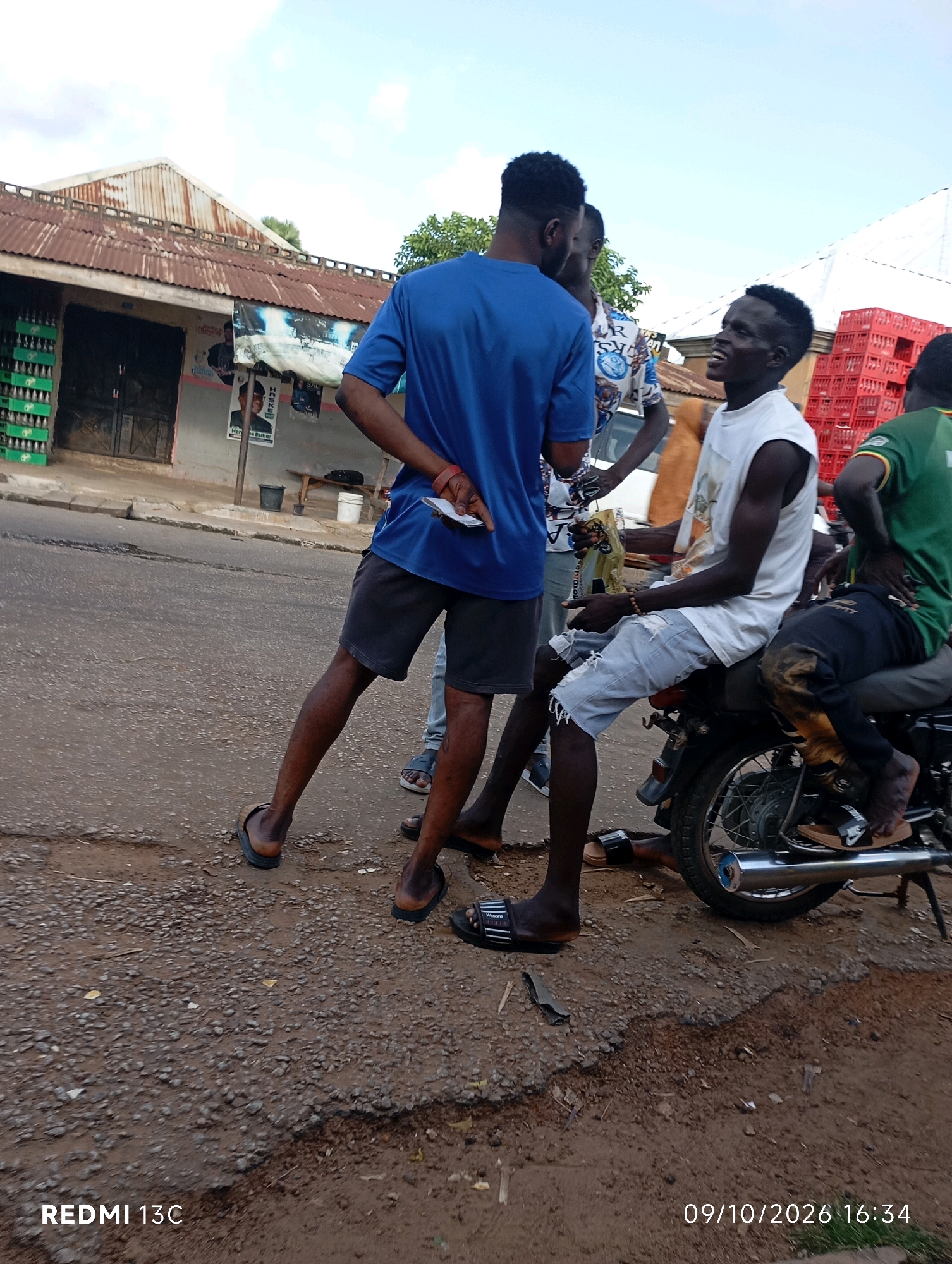The image captures a street scene in Shendam, Nigeria. In the foreground, four young men are interacting. One man, wearing a blue t-shirt and dark shorts, stands with his back mostly to the camera, holding a piece of paper. To his right, another man in a white tank top and light denim shorts sits on a motorcycle, looking to his left and talking. A third man, partially visible in a green shirt, sits behind him on the motorcycle. A fourth man is standing between the first two, his face obscured. The background features a building with a corrugated metal roof, stacks of green crates, and red plastic crates. The ground is a mix of asphalt and dirt, with some debris present. The sky is overcast. A timestamp in the lower right corner reads "09/10/2026 16:34", and "REDMI 13C" is in the lower left.