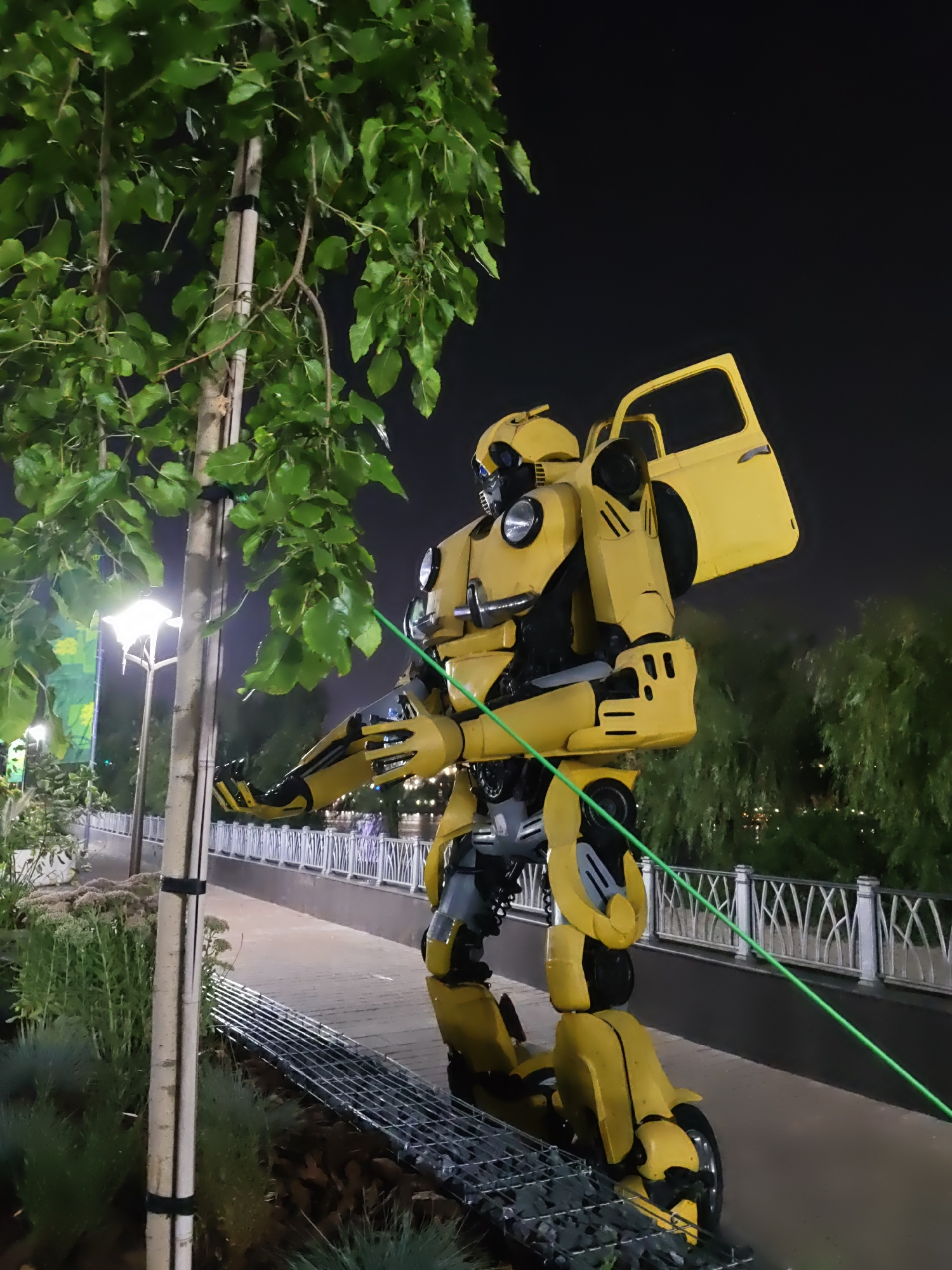 A life-sized statue of the Transformer character Bumblebee stands in an outdoor park at night. The statue is predominantly yellow with black accents and is posed in a dynamic stance, with its arms outstretched. A yellow car door is attached to its back, open and angled upwards. The statue is situated next to a tree with green leaves and is positioned on a paved walkway. In the background, there are streetlights, a white decorative fence, and lush green trees. A bright green laser line is visible, stretching diagonally across the image from the statue's chest towards the right. The scene is dimly lit, suggesting it is evening or nighttime.