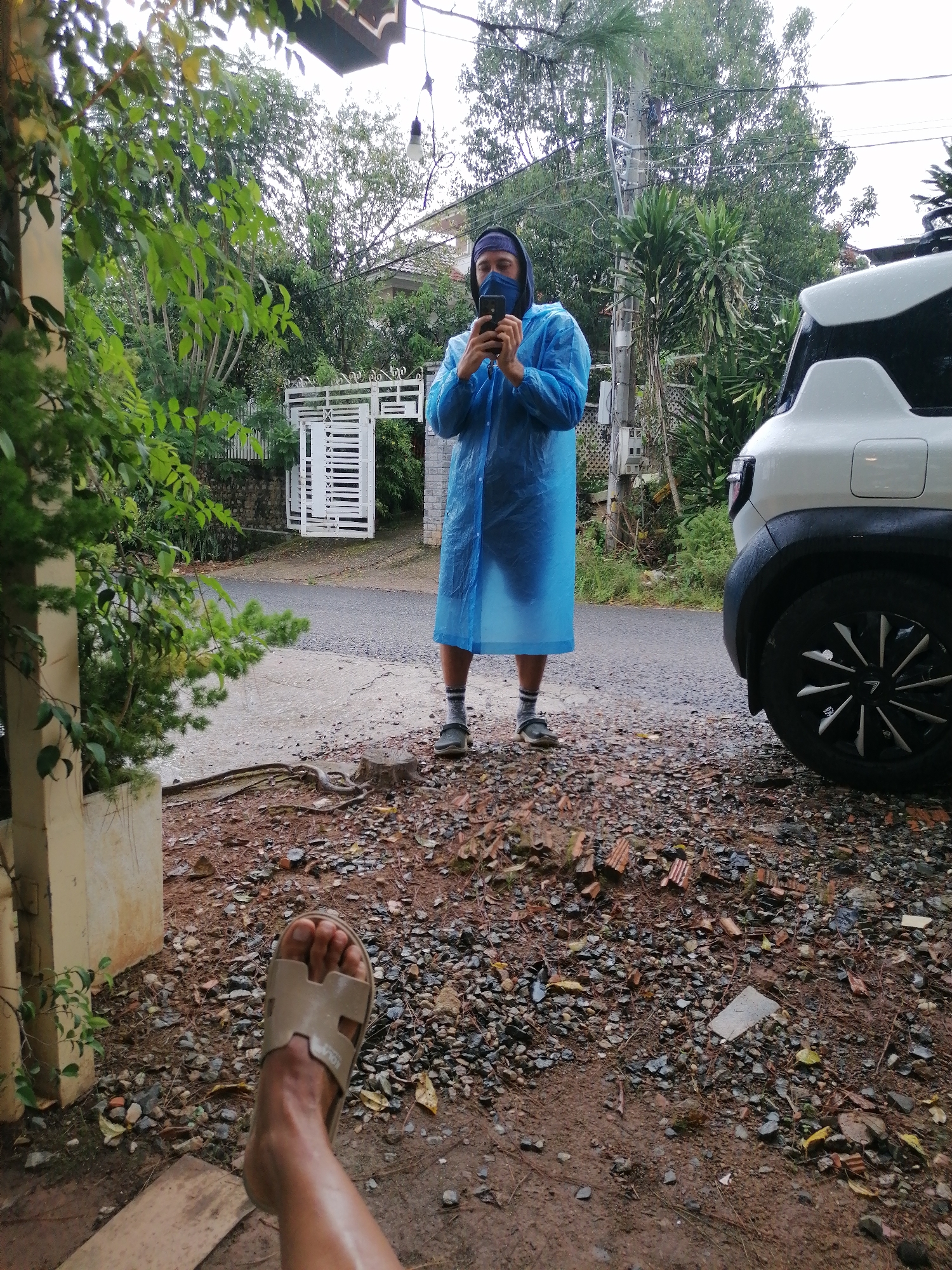 The image depicts an outdoor scene with a person wearing a blue rain poncho and a dark blue mask, holding a smartphone to take a selfie. They are standing on a wet, gravelly surface, with a white car parked to their right and lush green foliage in the background. A white gate and a residential building are visible further back. The foreground features a person's leg wearing a light brown sandal, suggesting a low-angle perspective. The environment appears damp, possibly indicating recent rainfall, consistent with the weather conditions often experienced in Lang Biang, Đà Lạt, Vietnam, known for its cool, humid climate and frequent showers. The overall impression is casual and candid, possibly capturing a moment during inclement weather.