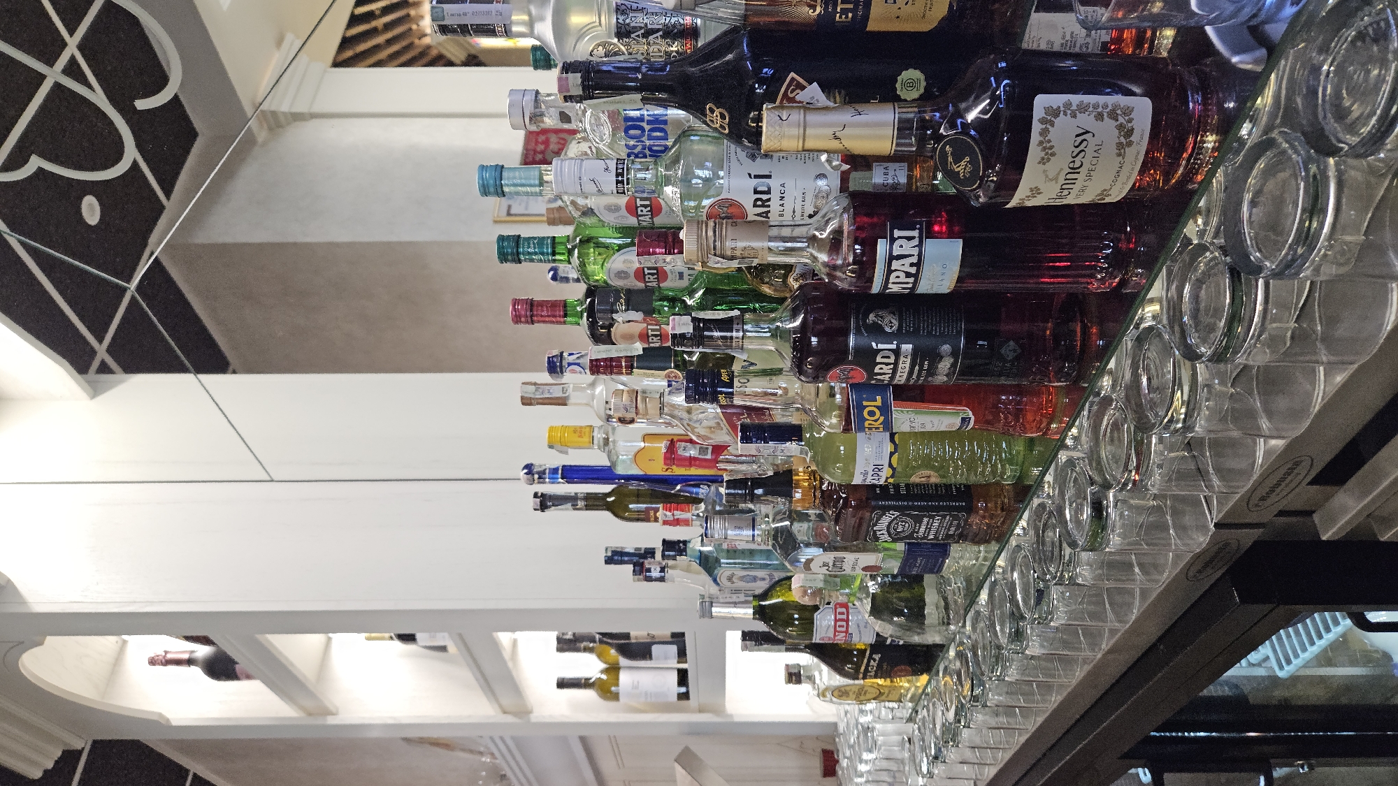 The image depicts a well-stocked bar, likely in a restaurant or establishment in Unknown, Unknown. The foreground is dominated by a row of clean, empty glasses, mostly tumblers and rocks glasses, arranged on a reflective surface, presumably a bar top. Behind the glasses, an extensive collection of liquor bottles is displayed on shelves and the bar top. The bottles are of various shapes, sizes, and colors, indicating a wide selection of spirits, liqueurs, and possibly wines. Brands such as Jack Daniel's, Hennessy, Absolut Vodka, and several Italian aperitifs like Campari and Aperol are identifiable.
In the background, white shelving units hold more bottles, and a mirrored wall reflects the bar and its contents, creating a sense of depth and expanding the visual space. The lighting appears to be ambient, highlighting the bottles and glassware. The overall impression is one of a professional and inviting bar setup, ready for service. There are no people visible in the image. The architectural style of the shelving and the mirrored elements suggest a modern or contemporary design. The ceiling features a patterned treatment.