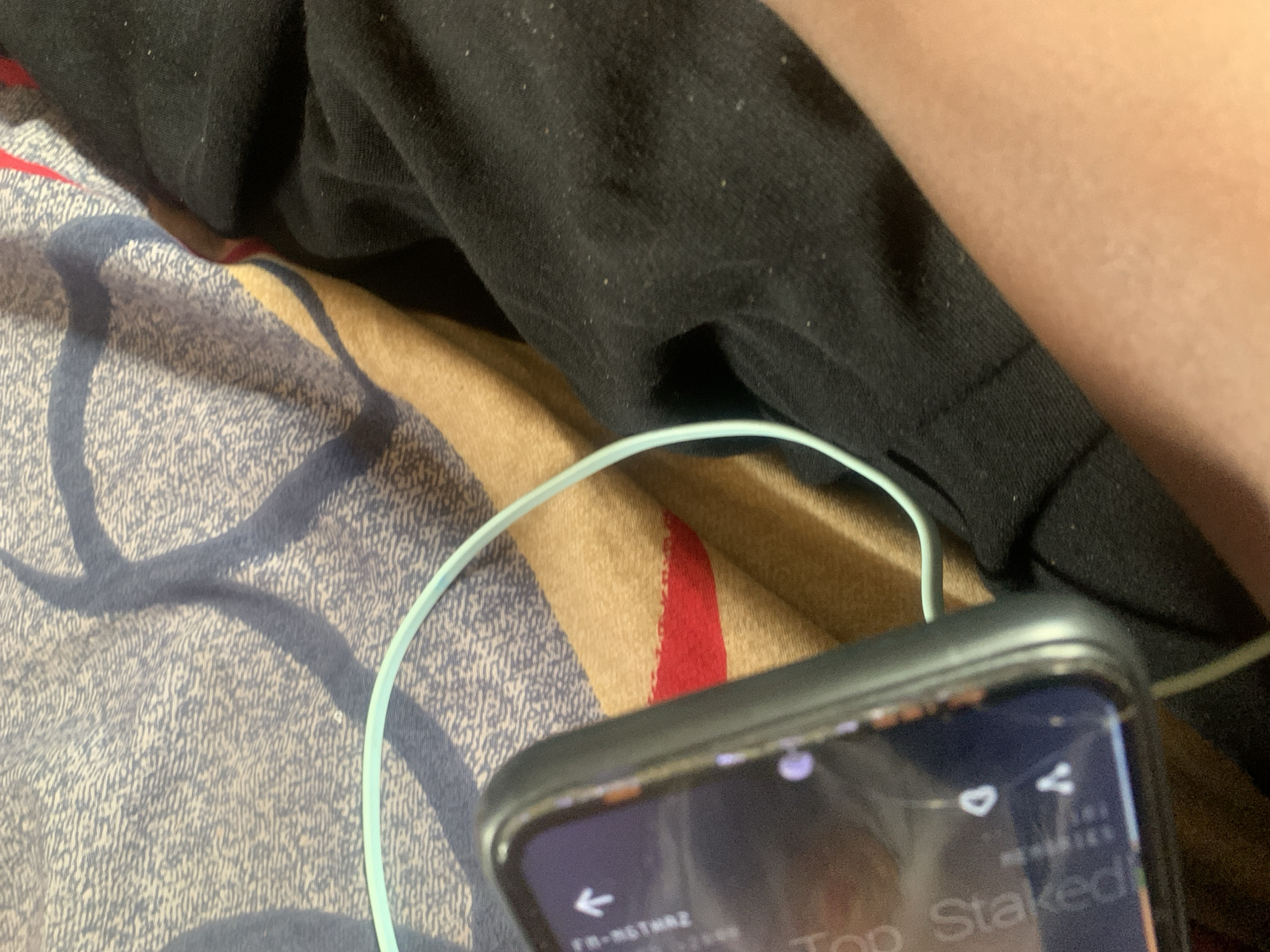 The image shows a close-up view of a person's arm and a mobile phone. The phone is partially visible on the left side of the frame, with its screen displaying some text and icons. A light blue cable is connected to the phone and extends out of view. The arm appears to be resting on a surface covered with a patterned fabric that includes blue and tan colors, with a red stripe. The overall scene suggests a casual, indoor setting. There is no information present in the image that indicates a specific location in Jauro Yinu, Nigeria.