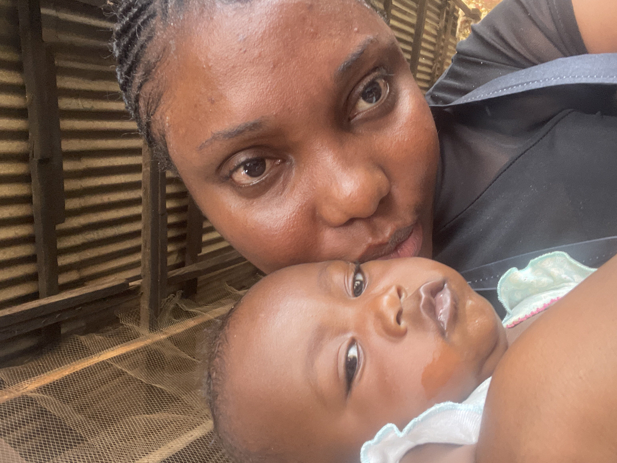 The image presents a warm, close-up portrait featuring an adult and a baby, likely a mother and child, captured in what appears to be a domestic or sheltered setting in Jos, Nigeria. The adult, with dark skin and braided hair, is positioned in the upper portion of the frame, looking directly towards the viewer with a tender expression and lips slightly puckered as if preparing for or having just given a kiss. She is wearing a dark-colored top.
The baby, also with dark skin, lies horizontally in the lower right section of the image, appearing to look upwards or towards the camera. There is visible drool or moisture around the baby's mouth and on the cheek. The baby is dressed in a light-colored garment, possibly a onesie, featuring ruffled edges with light blue/green and pink trim.
The background reveals corrugated metal sheeting, indicative of a wall or roof, interspersed with wooden beams and planks, suggesting a rustic or humble structure. A light-colored mesh material, resembling a mosquito net, is visible in the lower-left background. The natural lighting suggests the picture was taken during the day. The scene conveys an intimate moment of affection between the adult and the baby. No text is visible in the image.