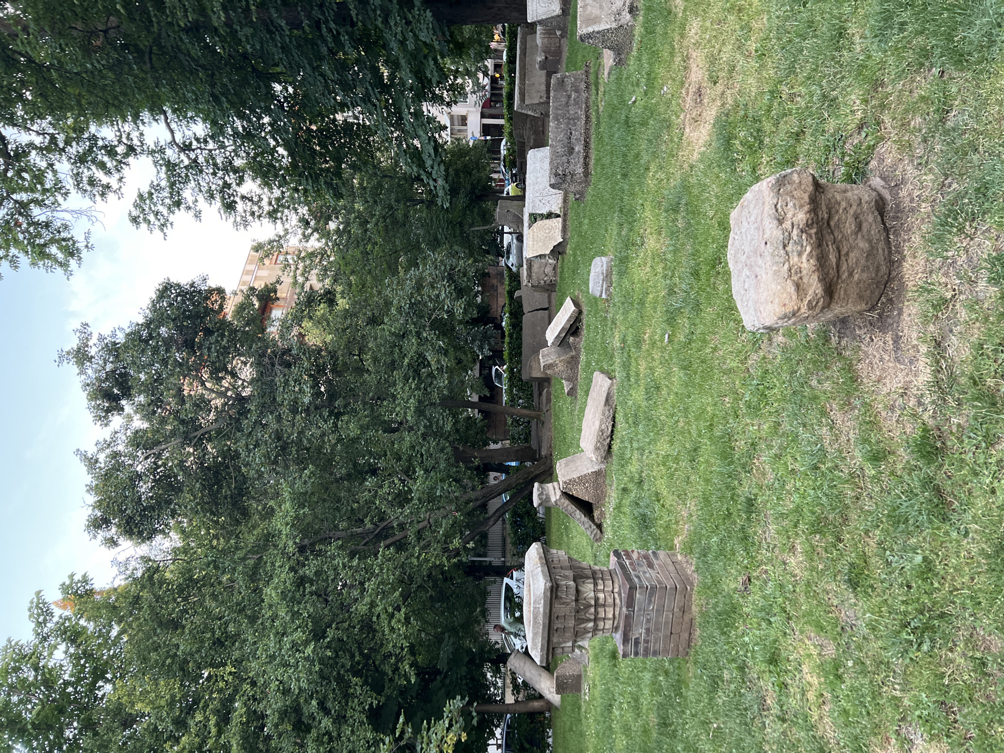 In the city of Sofia, Bulgaria, this image captures an outdoor scene depicting a collection of historical stone artifacts arranged on a grassy field. The artifacts appear to be remnants of ancient structures, including fragments of columns, bases, and inscribed stones, scattered across the lawn. In the foreground, a prominent stone pedestal with intricate carvings is visible, alongside other weathered stone blocks. The background features lush green trees and the facade of a multi-story building. Several vehicles are parked on a street adjacent to the field, and a few individuals can be observed in the distance. The sky above is partly cloudy. This scene is likely located in a museum or archaeological site, possibly the National Archaeological Institute with Museum in Sofia.