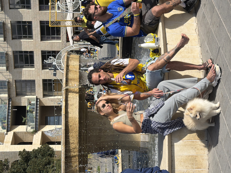 The image depicts an outdoor scene in a public plaza in Tel Aviv, likely within the Sarona Market area, identifiable by the distinctive modern light-colored stone buildings with balconies and large windows in the background. The foreground features three individuals seated on a light-colored stone ledge beside a flowing, multi-tiered stone fountain, under bright daylight.
On the left, a woman in her 20s-30s wears an olive green crop top, light grey wide-leg trousers, and sunglasses. A blue and white plaid shirt is tied around her waist. She holds a clear glass with a straw in her left hand, while her right hand is near her hair. A small, fluffy white Pomeranian-type dog is on a pink leash connected to her, standing on the pavement.
In the center, a man in his 30s-40s with dark dreadlocks and visible leg tattoos wears a yellow and white t-shirt featuring a blue 'P' logo and '5G' text, paired with ripped denim shorts. He is drinking from a brown bottle. His left foot is bare, resting on a white sneaker, while his right foot wears a flip-flop. A white insulated water bottle and a silver rectangular device are on the ledge beside him.
To the right, a man in his 20s-30s with a beard and sunglasses wears a blue t-shirt with a yellow crest and "ISRAEL" text, khaki shorts, and a camouflage-patterned cross-body bag. He holds a clear plastic cup. A yellow wristband is visible on his left wrist.
All three individuals are seated facing generally forward. In the background, numerous other people are visible, many also wearing yellow and blue attire, suggesting a public gathering. Decorative white metal archways, possibly illuminated, are also present further in the background.