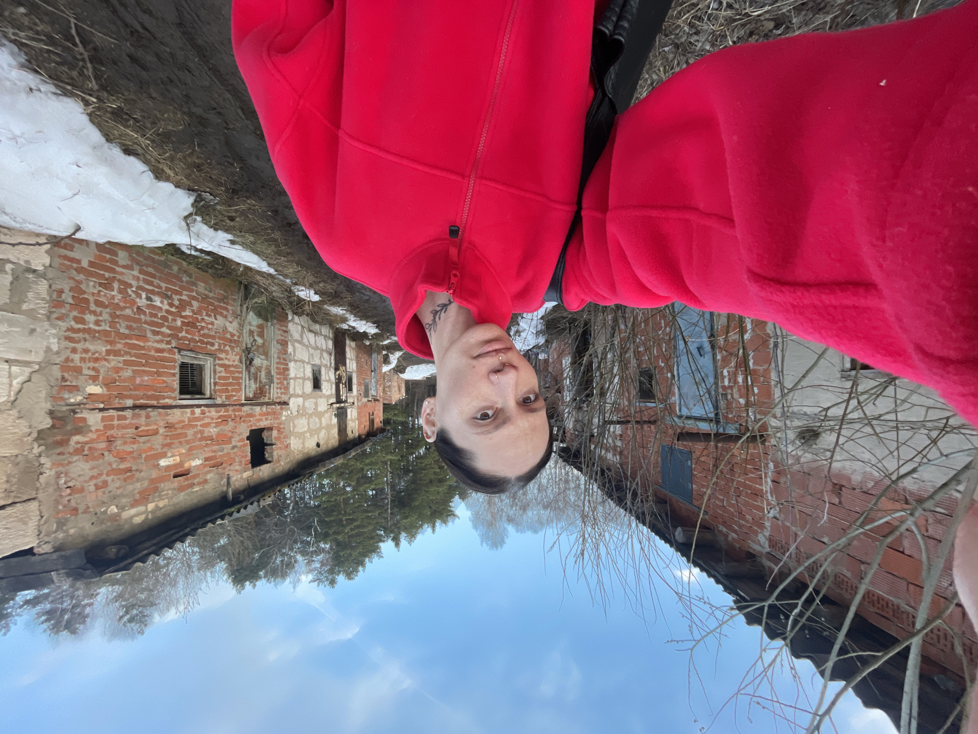 The image presents a person wearing a red fleece jacket, oriented with their head towards the bottom of the frame. The background reveals a dilapidated brick building, consistent with the architectural style found in Oktyabr'skii, Russia. Patches of snow are visible near the buildings, suggesting a winter or early spring setting. Bare trees are also in the background, against a blue sky with clouds. The buildings are in a state of disrepair, with missing sections and deteriorated brickwork.