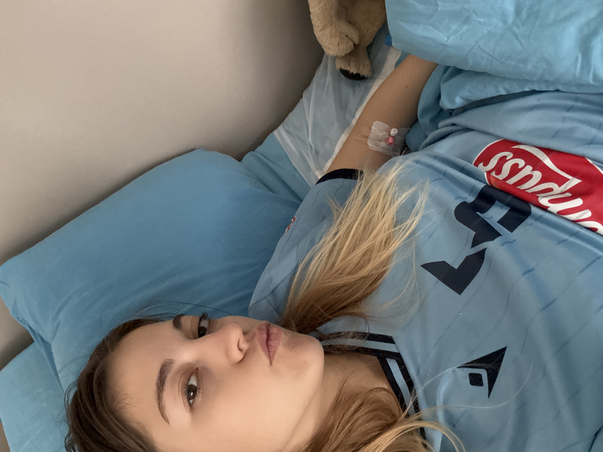 A woman is lying in a bed with blue sheets and a pillow. She has an IV in her arm. She is wearing a light blue soccer jersey. A stuffed animal is at the top of the frame. The location cannot be ascertained from the visual information provided.