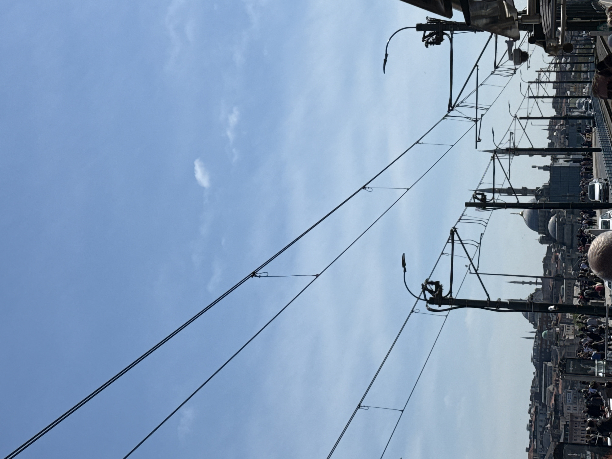 The image shows a view of a city skyline in Unknown, Unknown, possibly taken from a high vantage point or bridge. Overhead electrical wires with support structures stretch across the top portion of the frame against a blue sky with some clouds. The cityscape features buildings of varying heights and styles, including domed structures which indicate the presence of mosques or religious buildings. A crowd of people is visible along the edge of the structure from which the image is taken.