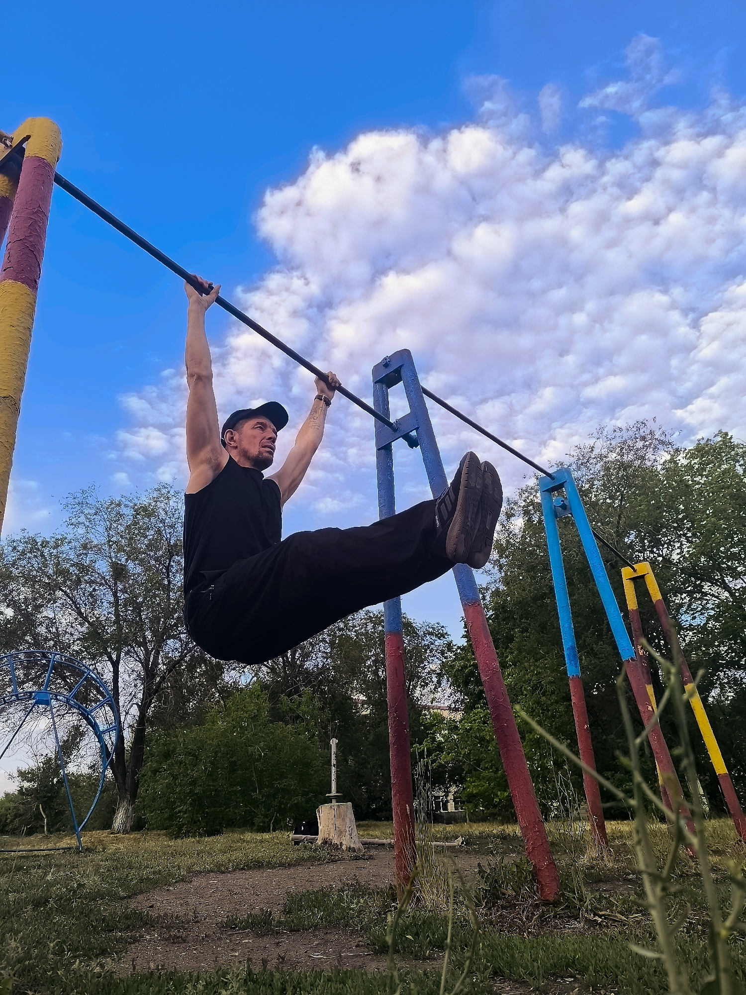An adult male, wearing a black sleeveless shirt, black pants, and a black baseball cap, is performing a leg raise exercise on a horizontal bar at an outdoor fitness equipment area. He is suspended from the bar with his arms extended and his legs held straight out in front of him, parallel to the ground. The fitness equipment consists of a horizontal bar supported by colorful red, blue, and yellow posts. In the background, there are trees, a partially visible blue metal structure, and a bright blue sky with scattered white clouds. The ground appears to be dirt and grass. The scene is outdoors in what appears to be a public park or recreational area, potentially within the city of Qarağandı, Kazakhstan.