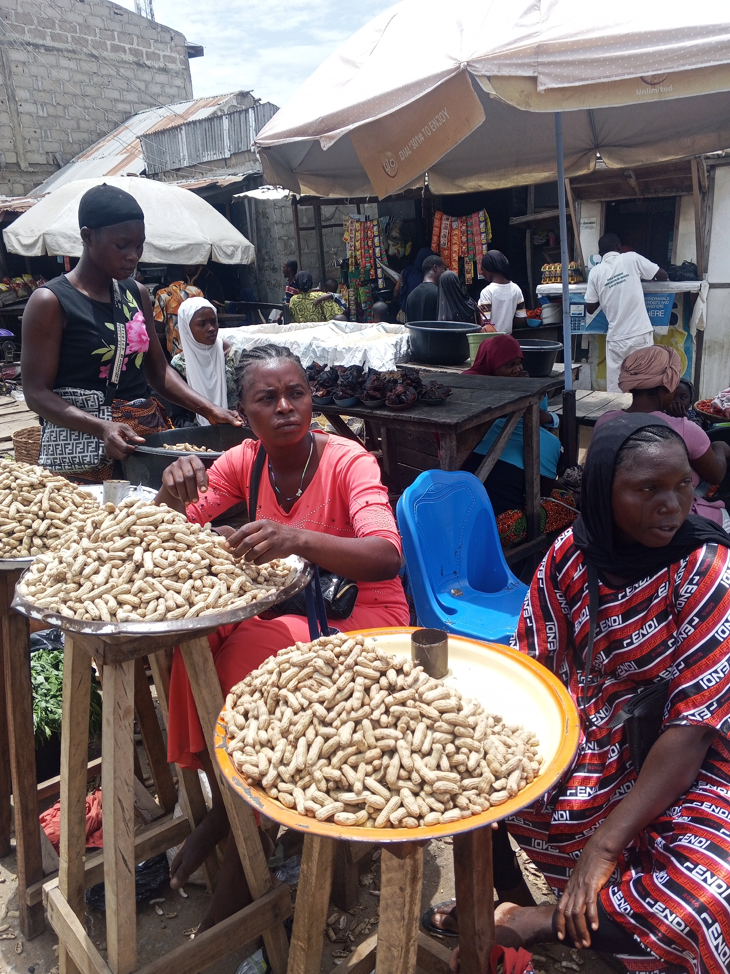 In Usunwe, Nigeria, a market scene unfolds under a bright, overcast sky. The foreground is dominated by three women and a vast quantity of unshelled peanuts. A woman in a black sleeveless top with a floral design stands to the left, her gaze directed downwards. Beside her, a woman in a vibrant coral-pink top is seated and actively sorting through a large metal tray filled with peanuts. Her focus is on the task at hand, her fingers delicately sifting through the nuts. To the right, another woman, also seated, wears a black headscarf and a distinctive red, black, and white striped garment emblazoned with a repeating logo pattern. She is looking slightly off to the side. The peanuts are displayed on large, shallow metal trays atop rustic wooden stands. Scattered peanut shells litter the ground beneath the stands, indicating recent activity. Behind the women, other market stalls and structures are visible, featuring various goods, umbrellas providing shade, and more people in the background, suggesting a busy marketplace. The overall impression is one of daily commerce and livelihood in a Nigerian town.