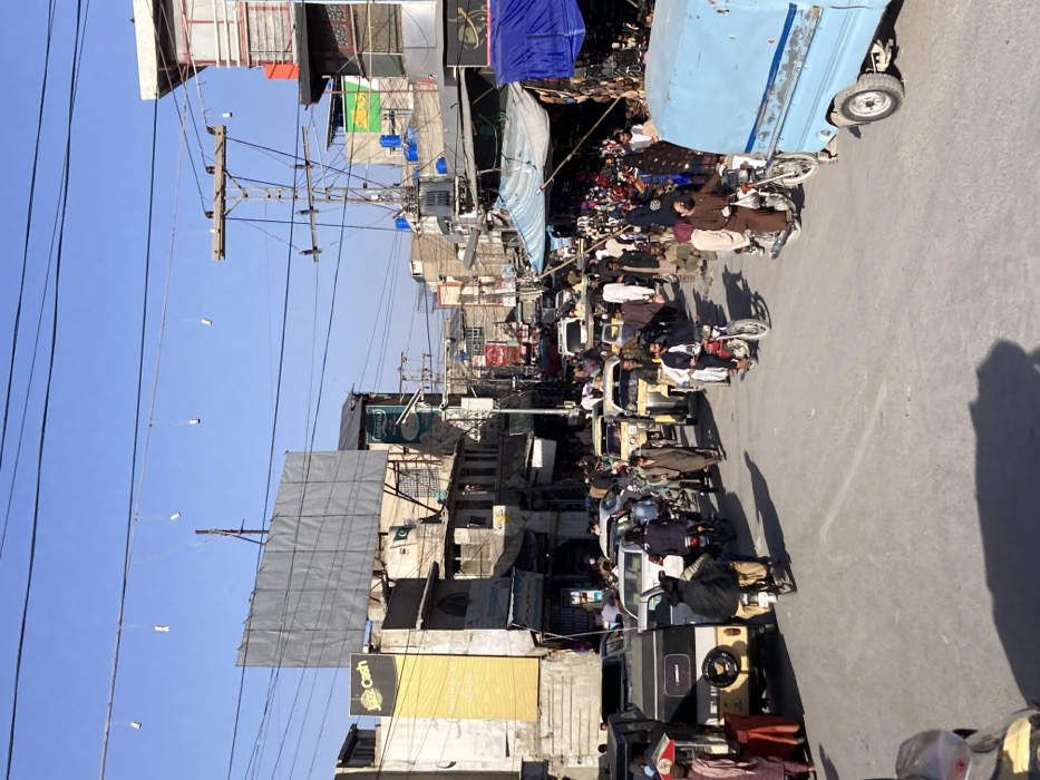 This image depicts a densely populated urban street in Quetta City Tehsil, Pakistan, under a clear blue sky. The street is a central thoroughfare characterized by heavy mixed traffic and pedestrian activity.
**Environment:** The paved road is lined with multi-story concrete and brick buildings, many appearing weathered or unfinished. Extensive networks of overhead power lines and communication cables crisscross the sky, supported by numerous wooden utility poles, some equipped with transformers. On the left side, a large, blank grey billboard is mounted on a building, below which a prominent yellow vertical banner advertises "Jazz Cash." Further down, a Pakistani flag is visible on another building. The right side of the street features numerous market stalls operating beneath blue and light-colored tarpaulin covers, displaying various goods. Small, decorative string lights are suspended high above the street between buildings.
**People:** A substantial crowd of individuals, predominantly adult males dressed in traditional attire such as shalwar kameez, some wearing turbans or caps, occupies the sidewalks and intermingles with vehicles in the street.
**Vehicles:** Vehicular traffic is heavy and appears congested. It comprises numerous motorcycles, including red and black models, with riders and some passengers. Several yellow-bodied auto-rickshaws are present. A variety of cars, including a silver sedan and a black SUV, are visible in the flow of traffic. In the lower right foreground, a light blue, small utility truck with an open cargo bed is parked or moving slowly.
**Actions and Interactions:** Pedestrians are walking, standing, and navigating the crowded environment. Individuals are operating motorcycles and other vehicles within the congested street. Commercial activity is evident from the numerous market stalls and shopfronts. The scene indicates a busy daytime commercial and transit hub.