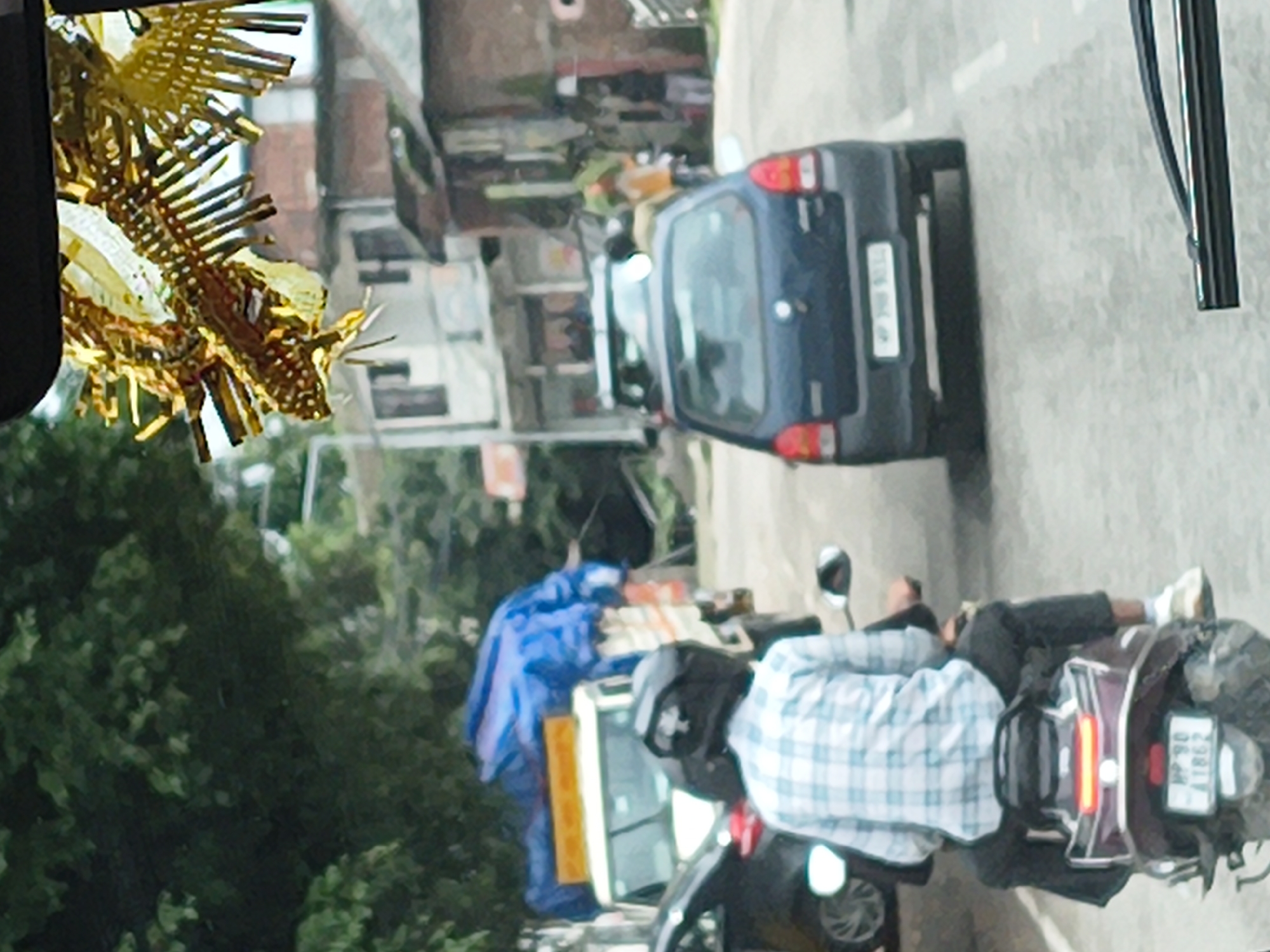 The image shows a street scene in Sidhpur, India, with a focus on two people on a motorcycle. The motorcycle is dark colored with a maroon seat and has a license plate that reads "MP 09 Z 7886". One person is wearing a blue and white plaid shirt and a black helmet, and is seated on the back of the motorcycle. The other person is partially visible, wearing dark pants and light-colored shoes.
Behind the motorcycle, a blue tarp covers what appears to be some goods or equipment. To the left of the motorcycle, a dark blue car is parked on the street. The car has a license plate that is partially visible. Further in the background, buildings with multiple stories are visible, suggesting a semi-urban or town setting. The overall impression is of a street scene, possibly during daylight, with everyday activities taking place. There are trees and greenery in the background, indicating an outdoor setting. A gold, spiky decoration is visible on the left side of the image, partially obscuring the view of the buildings. The image is taken from a slightly elevated perspective, looking down on the scene.