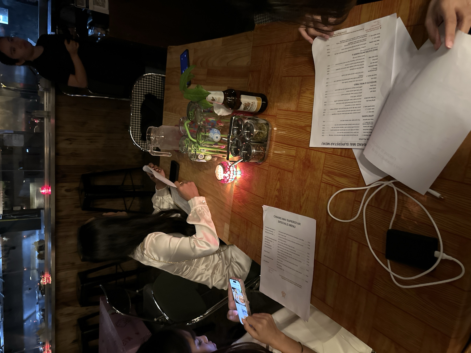 This image depicts a dimly lit indoor dining area, likely a restaurant, with a focus on a wooden table set for a meal. The atmosphere appears casual and intimate, with soft lighting emanating from a decorative candle. The background reveals a bar area with stainless steel surfaces and wooden paneling, suggesting a modern yet rustic aesthetic. The presence of menus, drinks, and condiments implies that the patrons are in the process of ordering or dining. The overall scene captures a moment of conviviality and relaxation, hinting at a pleasant dining experience.