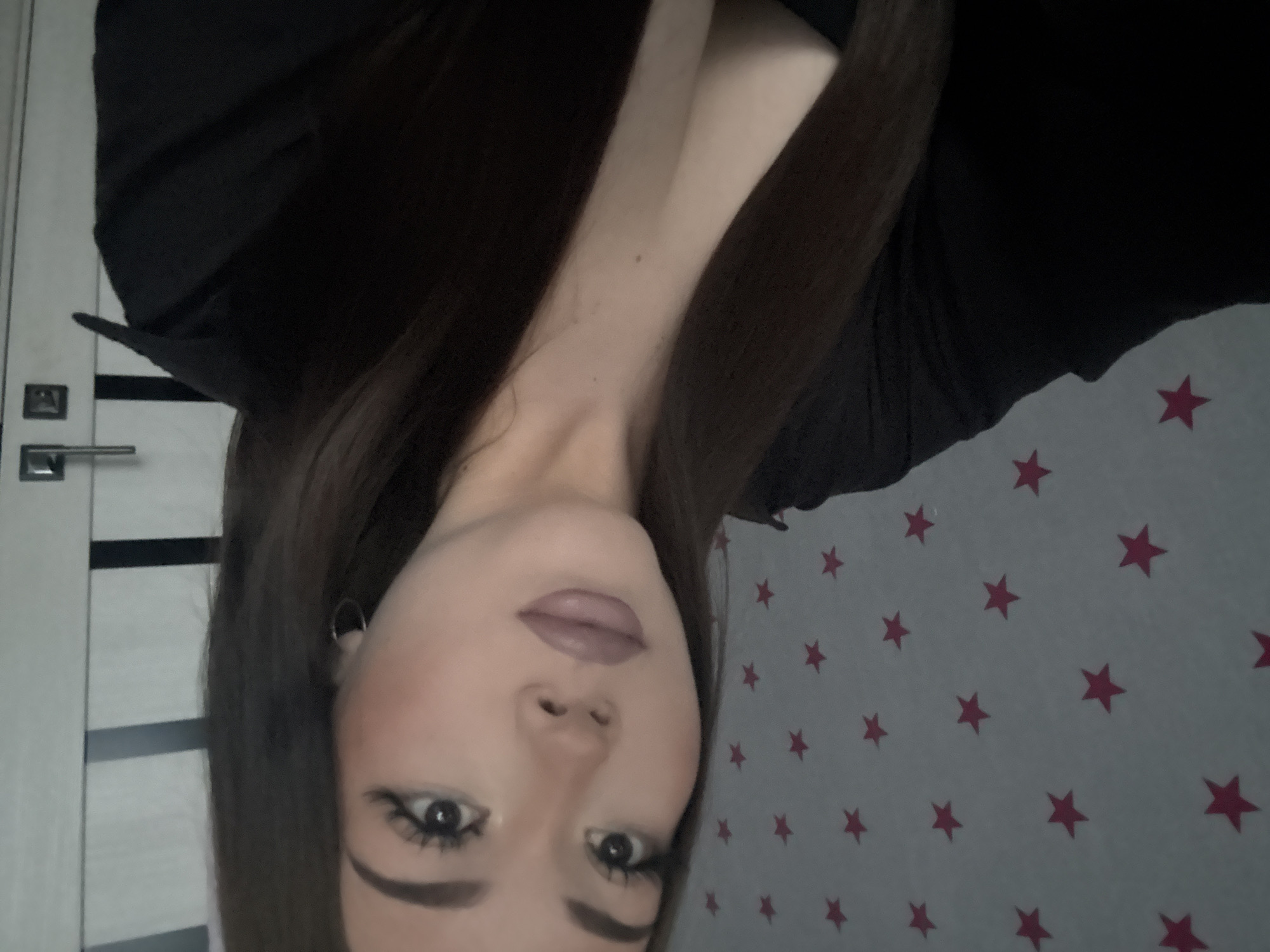 A medium shot shows a young woman with long, dark hair and fair skin, wearing a low-cut black top, indoors. Her makeup includes dramatic eyeliner and a muted lipstick. She is positioned in front of a light-colored wall adorned with pink stars. To her right, a white door with a silver handle is visible. The scene appears to be in a residential setting, and there are no explicit indicators placing it in Naberezhnye Chelny, Russia.
