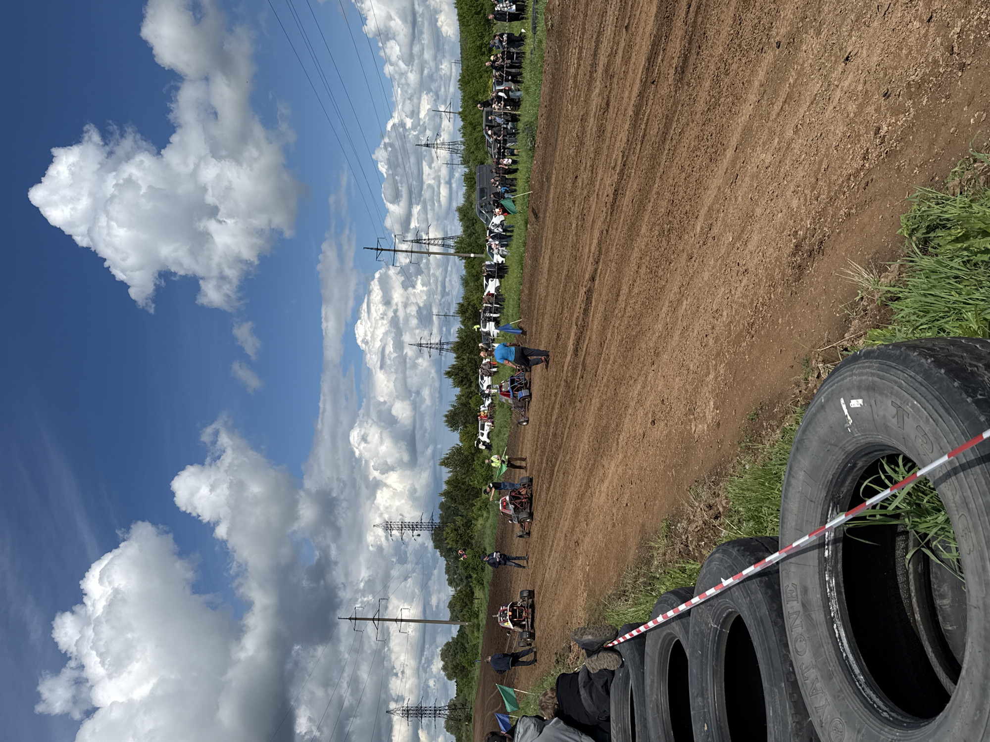 The image shows an outdoor race event in Votkinsk, Russia. The track is dirt, lined with tires and red-and-white striped tape. Several go-karts and race cars are present on the track, with a small crowd observing from a distance. Power lines are visible in the background against a partly cloudy sky. People are standing around the vehicles. Green vegetation surrounds the race area.