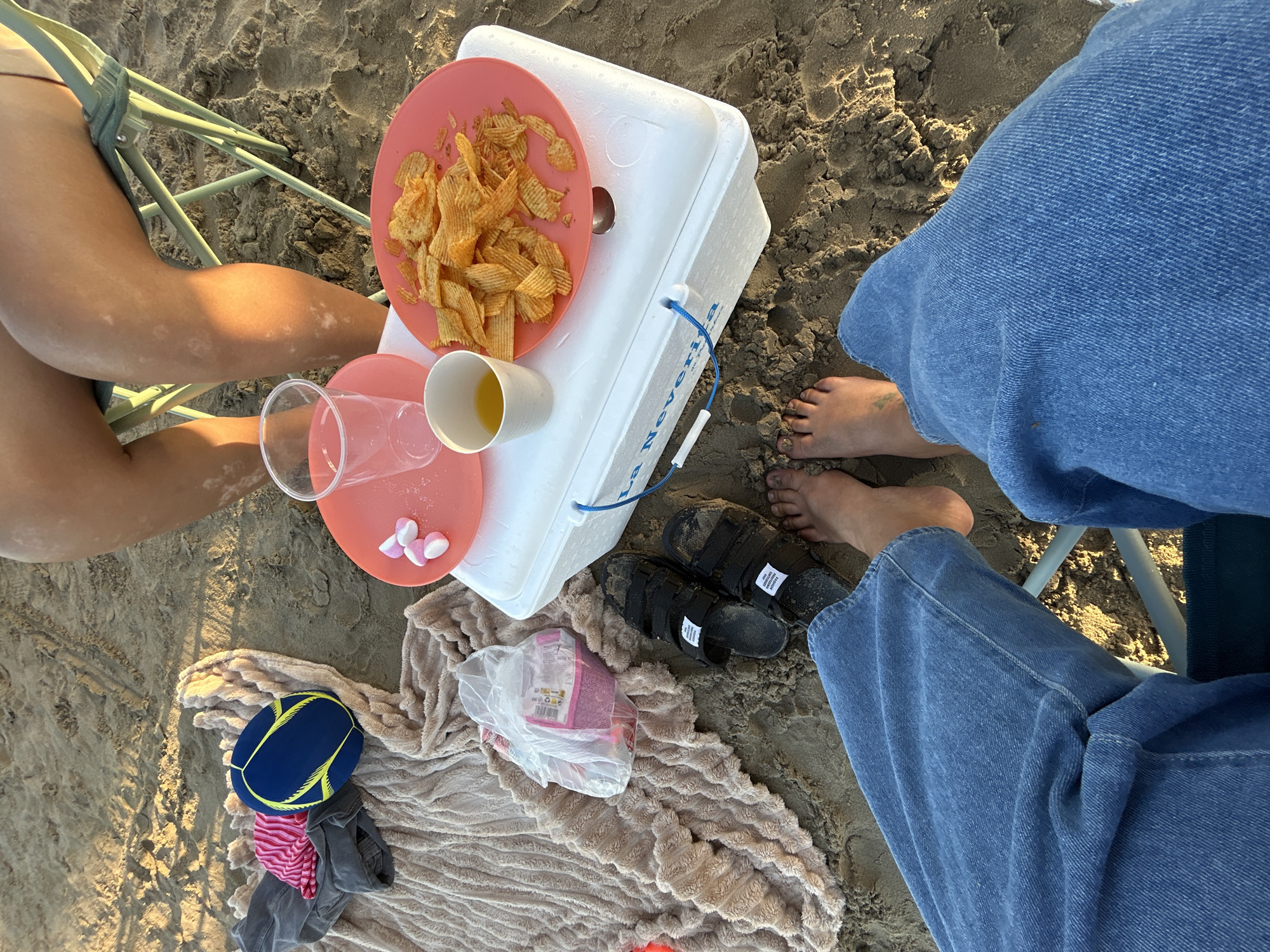 The image shows a close-up view of a beach scene in Castelló de la Plana, Spain, with two people sitting on chairs. The sand is visible, and the lighting suggests it is either early morning or late afternoon.
The foreground is dominated by the legs of two individuals. One person's legs are in the lower right of the frame, wearing blue denim jeans. Their bare feet, with visible toenails and some sand, are also present. The other person's legs are on the left, also bare and showing some tan lines. This person is sitting in a light-colored folding chair.
On the sand, between the two people, are various objects. A white cooler is prominently featured. On top of the cooler, there's a pink plate filled with ruffled potato chips. Next to it, a white cup holds a yellow liquid, likely a beverage. There are also some pink and white heart-shaped candies on a separate pink dish.
Below the cooler, a beige, fuzzy blanket is spread on the sand. On the blanket, a plastic bag contains more pink items, possibly snacks or candy. A blue and yellow disc, possibly a frisbee or a child's toy, is placed on top of some folded clothing, including a pink and white striped item and a grey garment.
Towards the upper right of the image, a pair of black sandals with velcro straps are visible, also covered in sand.
The overall impression is a relaxed beach outing, likely a picnic or a break during a day at the beach. The presence of snacks, drinks, and leisure items suggests a casual and enjoyable experience. The text "La Nevera" is visible on the side of the white cooler.