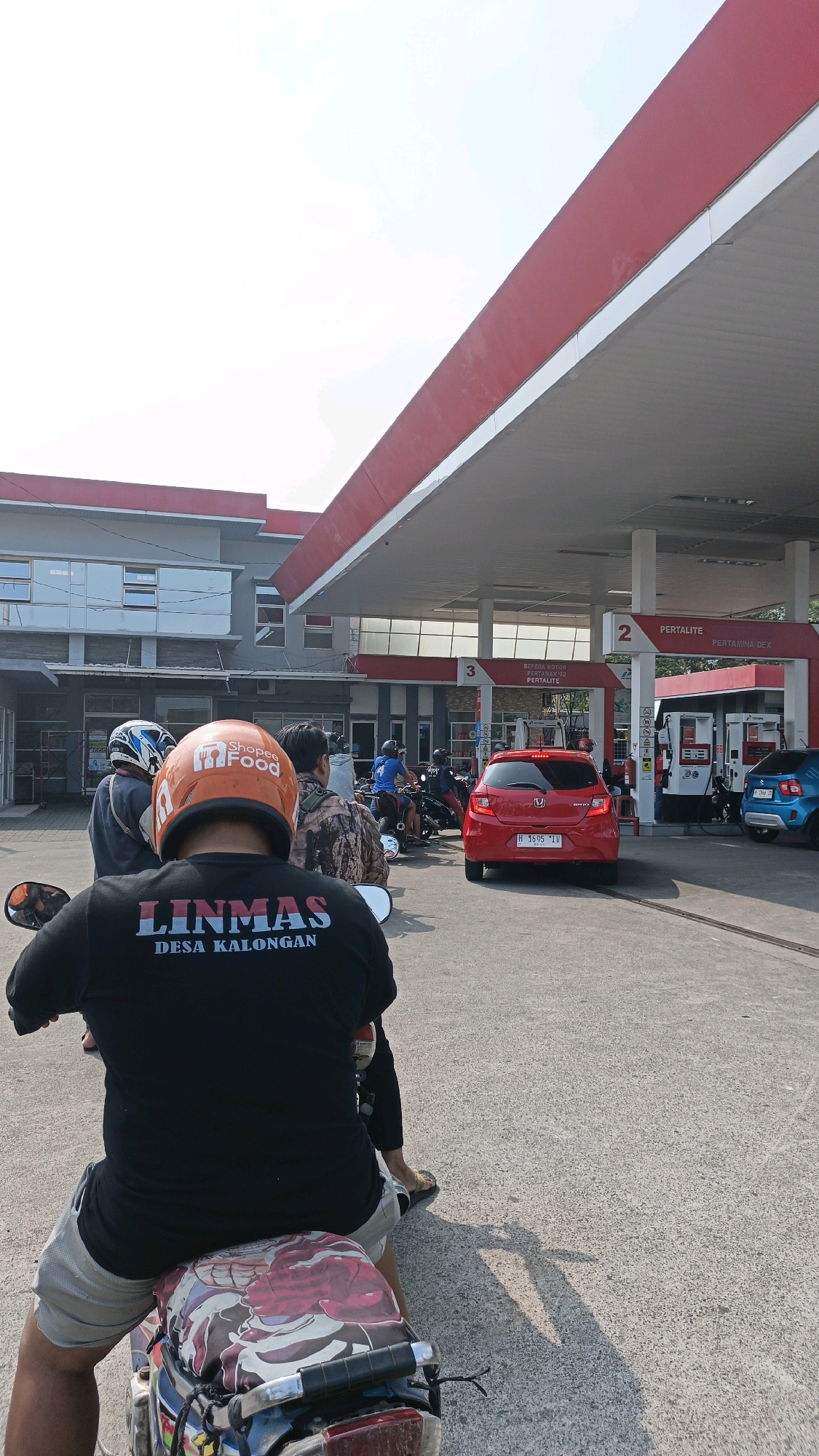 The image captures a scene at a gas station in Gedanganak, Indonesia, on a sunny day. In the foreground, a person is seated on a motorcycle, facing away from the camera. They are wearing a black t-shirt with white text that reads "LINMAS DESA KALONGAN" and light-colored shorts. The motorcycle's seat has a colorful cover.
Behind the person, several other motorcycles and a red car are visible, suggesting a queue or activity at the gas station. The gas station has a prominent red and white awning. Several gas pumps are present, with signage indicating fuel types like "PERTAMAX" and "PERTAMINA DEX." To the left of the gas station, a building with several windows is visible. The overall environment appears to be an outdoor urban setting.
