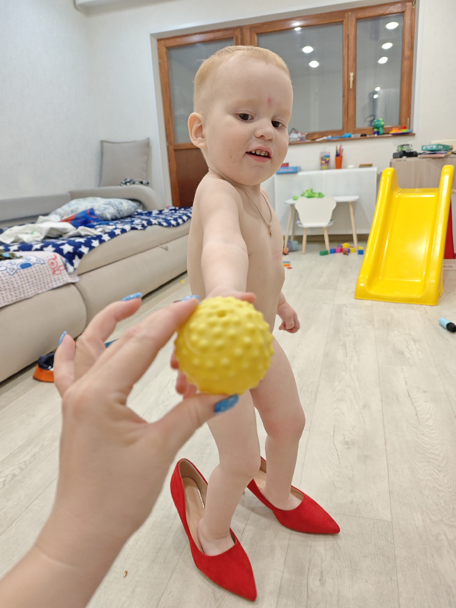 A close-up full shot in Markovskii, Russia, shows a fair-skinned toddler with red hair and a slight birthmark on their forehead standing on a light-colored wooden floor. They are nude, except for a gold necklace and red high-heeled shoes. The child is holding out a yellow ball towards the viewer. A fair-skinned adult hand with blue nail polish is also holding the ball. In the background, there is a beige sofa with a blue star-patterned blanket, a window with light filtering through, a yellow slide, and a white children's table with a white chair. Toys and crayons are also visible on a ledge near the window.