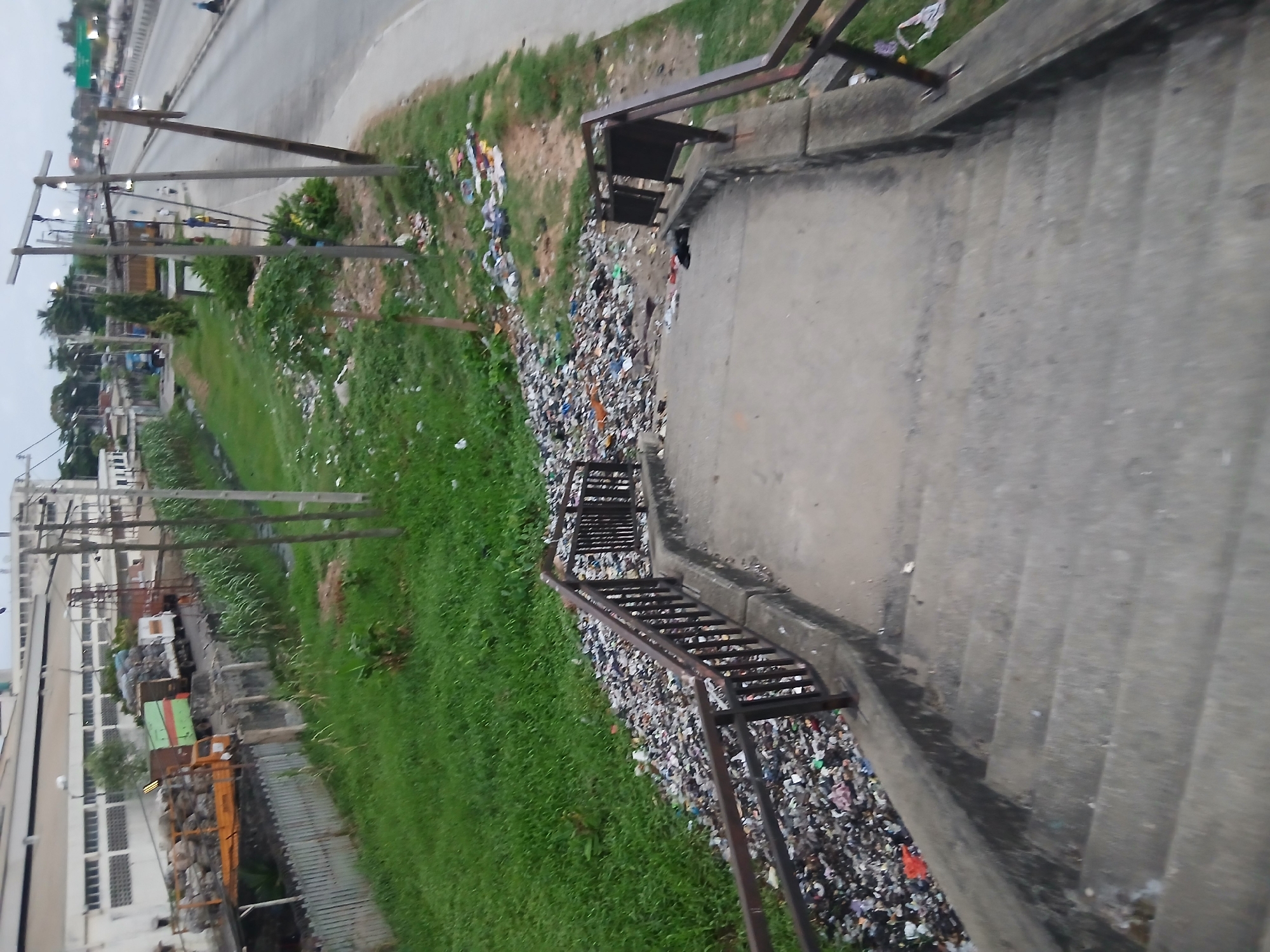 The image shows a view from the top of a set of concrete steps. The steps lead down to a grassy area littered with trash. A metal railing runs along the side of the steps. Across the grass is a street with vehicles, utility poles, and buildings. The image appears to be taken in an urban environment.