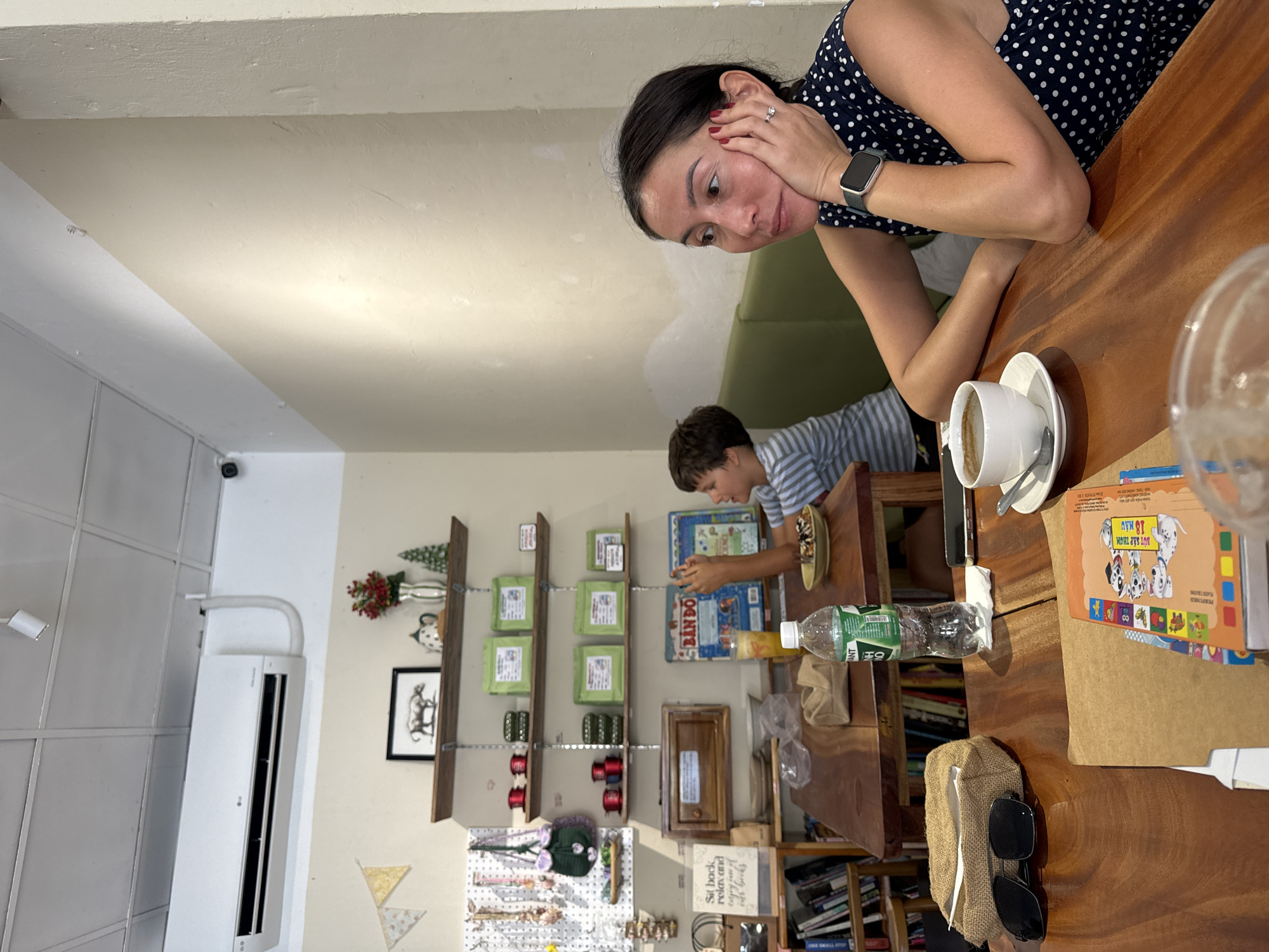 This image captures a casual indoor scene, likely in a cafe or a cozy shop, featuring a woman and a young boy. The photo is rotated, showing subjects at an angle.
In the foreground, a woman with dark hair, wearing a dark blue polka-dot top, rests her head on her hand, looking downwards towards the boy. She has a ring on her left ring finger and a smartwatch on her left wrist. Her expression appears thoughtful or pensive. Seated across from her at a wooden table is a young boy with dark hair, wearing a striped shirt. He is engrossed in an activity, possibly a board game or craft, with a small dish of food nearby.
The wooden table is laden with various items: a white cup of coffee or tea on a saucer with a spoon, a plastic bottle of Aquafina water, an empty glass, a pair of sunglasses in a burlap-like pouch, and a children's book featuring cartoon Dalmatians with the Vietnamese text "BÚT SÁP MÀU 18 MÀU" (Crayons 18 colors). A large "BINGO" board game box is also visible near the boy.
The background reveals the interior of the establishment, adorned with wooden shelves holding various products like green packaged goods (possibly "Trà Xanh" or Green Tea), small decorative items such as miniature evergreen trees and vases with red flowers, and a framed picture of a bovine animal. A pegboard display with accessories and a sign that reads "Sit back, relax and enjoy our books" suggest a relaxed atmosphere. A white air conditioning unit is mounted on the wall, and bookshelves filled with books are visible towards the bottom right. A security camera is also discernible near the ceiling.
The time of day appears to be daytime, given the general lighting. The scene conveys a quiet, relaxed moment between the two individuals in a quaint, perhaps artisan or book-themed, cafe setting in Phu Thuy Ward, Vietnam, as indicated by the visible Vietnamese text.