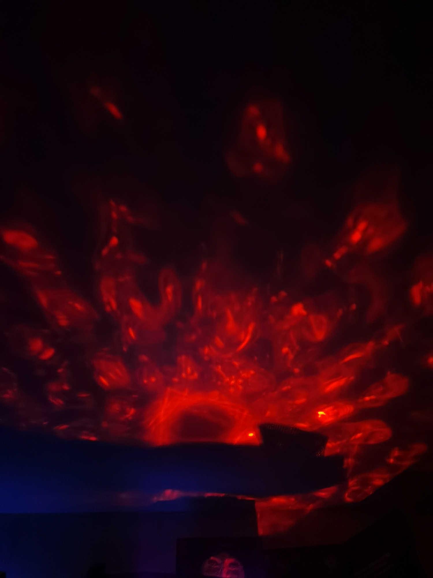 The image shows a dark room with red light patterns projected on the ceiling. The source of the light appears to be a device placed on a surface, creating the abstract, fiery-looking patterns above. There are no people visible. The environment is generally dark, with some objects silhouetted in the foreground. There is no indication this photo was taken in Mozhga, Russia, or any other specific location.