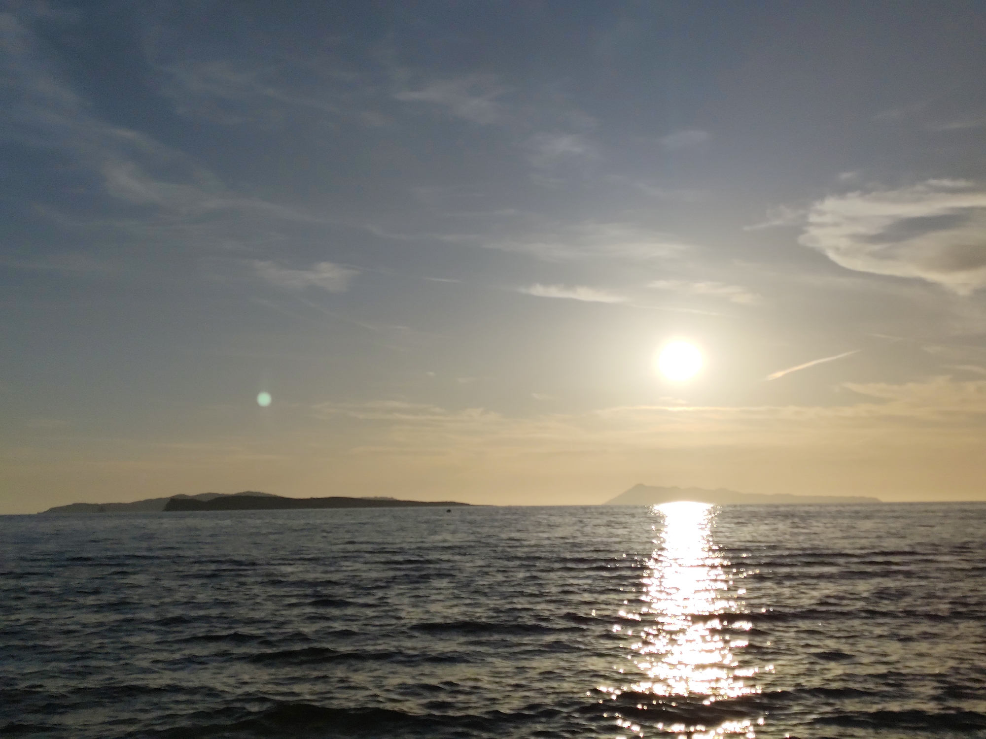 This image captures a seascape at either sunrise or sunset in Agios Stefanos, Greece. The horizon line is positioned approximately at the image's midpoint, dividing the frame into a sky upper half and a sea lower half.
The sky exhibits a gradient of muted blue-gray tones, transitioning to a pale yellow-orange near the horizon due to the sun's presence. Scattered, wispy white clouds are visible across the sky. A bright, circular sun is prominent in the mid-right portion of the sky, casting a strong glare. A distinct green circular lens flare is present in the upper left quadrant, and a faint contrail crosses the sky in the upper right.
The sea in the foreground is dark blue-gray with a rippled, textured surface indicating small waves. A brilliant, shimmering path of sunlight reflects across the water, originating from the sun and extending towards the viewer, creating numerous bright specular highlights on the water's surface.
Two distinct landmasses are visible on the distant horizon. The landmass to the left is lower and extends horizontally, while the one to the right appears higher and more mountainous, both silhouetted against the brighter sky. An unidentifiable small, dark object is faintly discernible on the water's surface between the left landmass and the sun's reflection path. No people, discernible boats, or other prominent man-made structures are present in the foreground or midground.