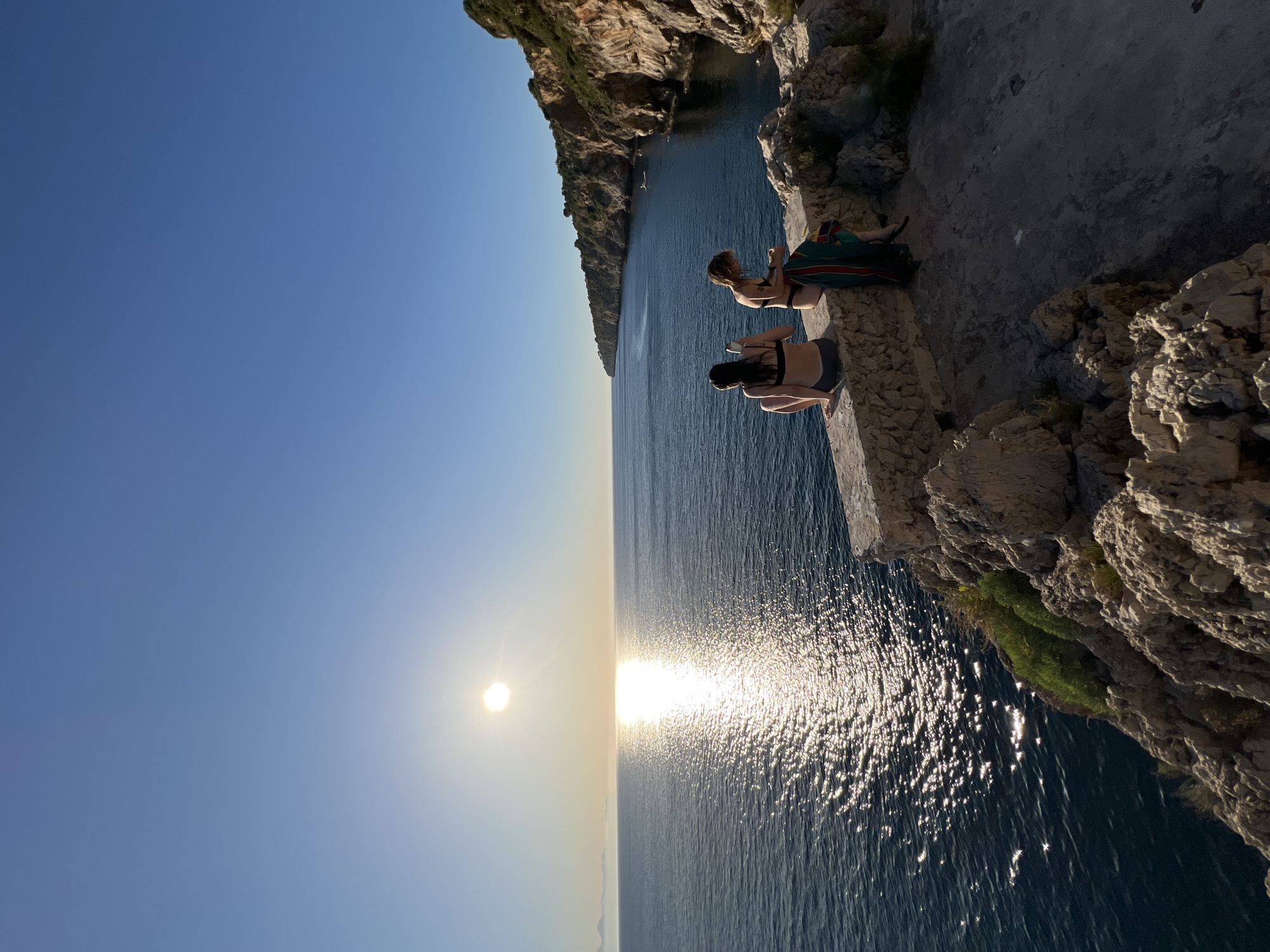 The image depicts a serene coastal scene at sunset in Terrasini, Italy. Two individuals, both wearing swimwear, are seated on a stone ledge overlooking the Mediterranean Sea. The sun, low on the horizon, casts a shimmering reflection on the water. The coastline is characterized by rugged cliffs adorned with vegetation, leading into the distance. One person on the left is holding a phone, while the person on the right appears to be tending to a towel. The environment is bathed in the warm, golden light of the setting sun, creating a peaceful and picturesque atmosphere.