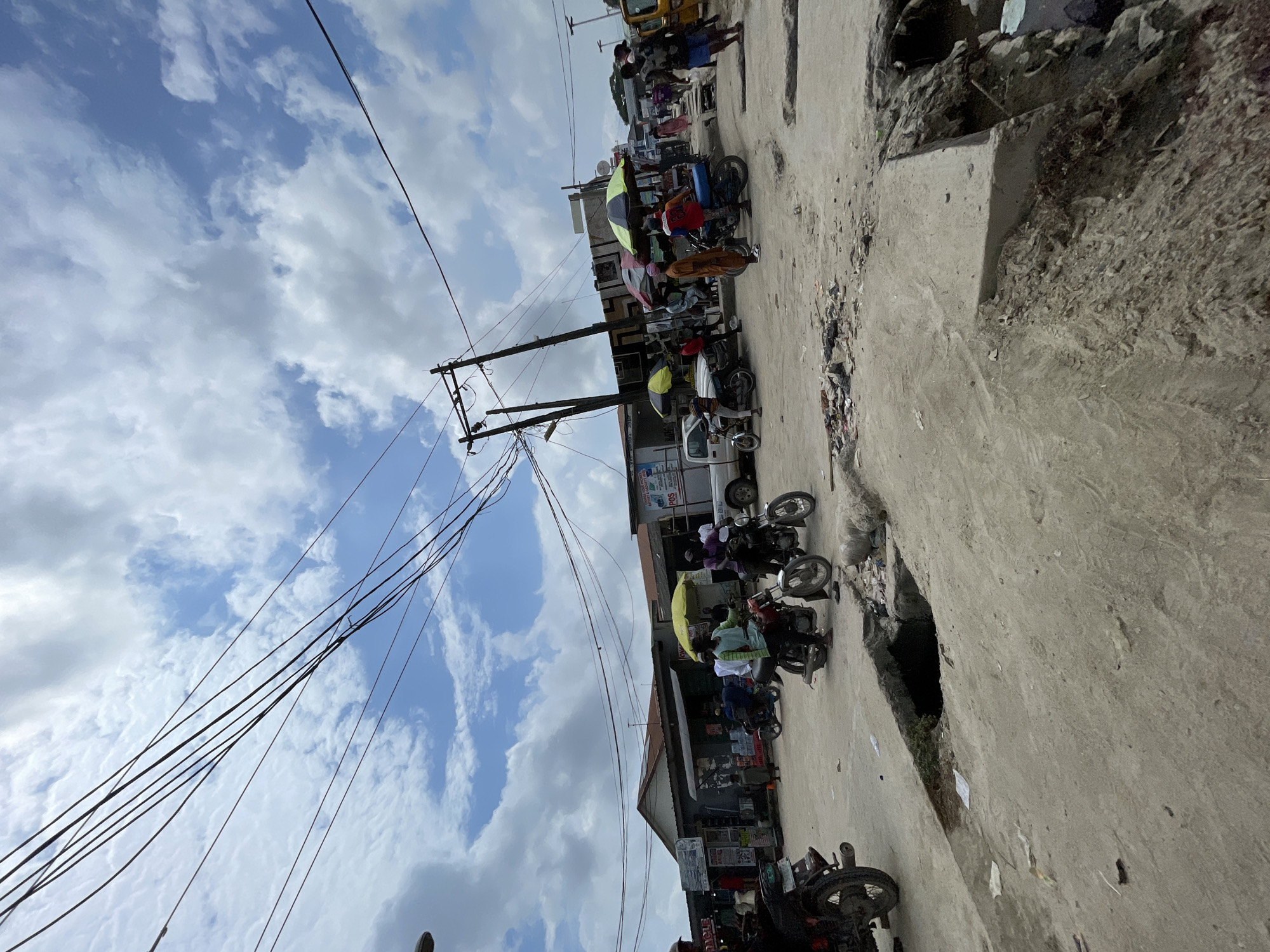 The image depicts a bustling street scene in Lagos, Nigeria, with a prominent focus on the urban environment and the daily activities of its inhabitants. The foreground is dominated by a cracked and uneven concrete road, with a large, dark opening suggesting a damaged drain or sinkhole. Debris is scattered across the ground, indicative of ongoing development or wear and tear.
In the mid-ground, motorcycles, a common mode of transport in Lagos, are parked and in motion. Several individuals are seen riding or standing near their motorcycles. Various shops and stalls line the street, with awnings and umbrellas providing shade. Some of these establishments display signage, although the text is largely illegible. People are casually dressed and engaged in various activities, suggesting a marketplace or commercial area. The presence of umbrellas, some with bright yellow and green colors, adds a splash of vibrancy to the scene.
The background reveals more buildings, utility poles with a dense network of overhead cables, and a truck. The sky is a mix of blue with scattered white clouds, indicating daylight. The overall impression is one of a dynamic and active urban setting, characteristic of many parts of Lagos, Nigeria. The scene is candid and captures the raw, unadulterated essence of street life in the city.